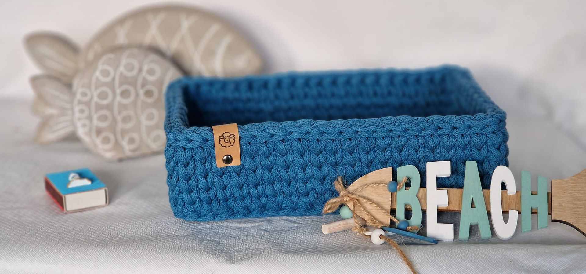 Blauer gehäkelter Korb mit dem Schild „Strand“. Weitere Dekorationen: ein Fisch, Streichhölzer und Bindfaden.