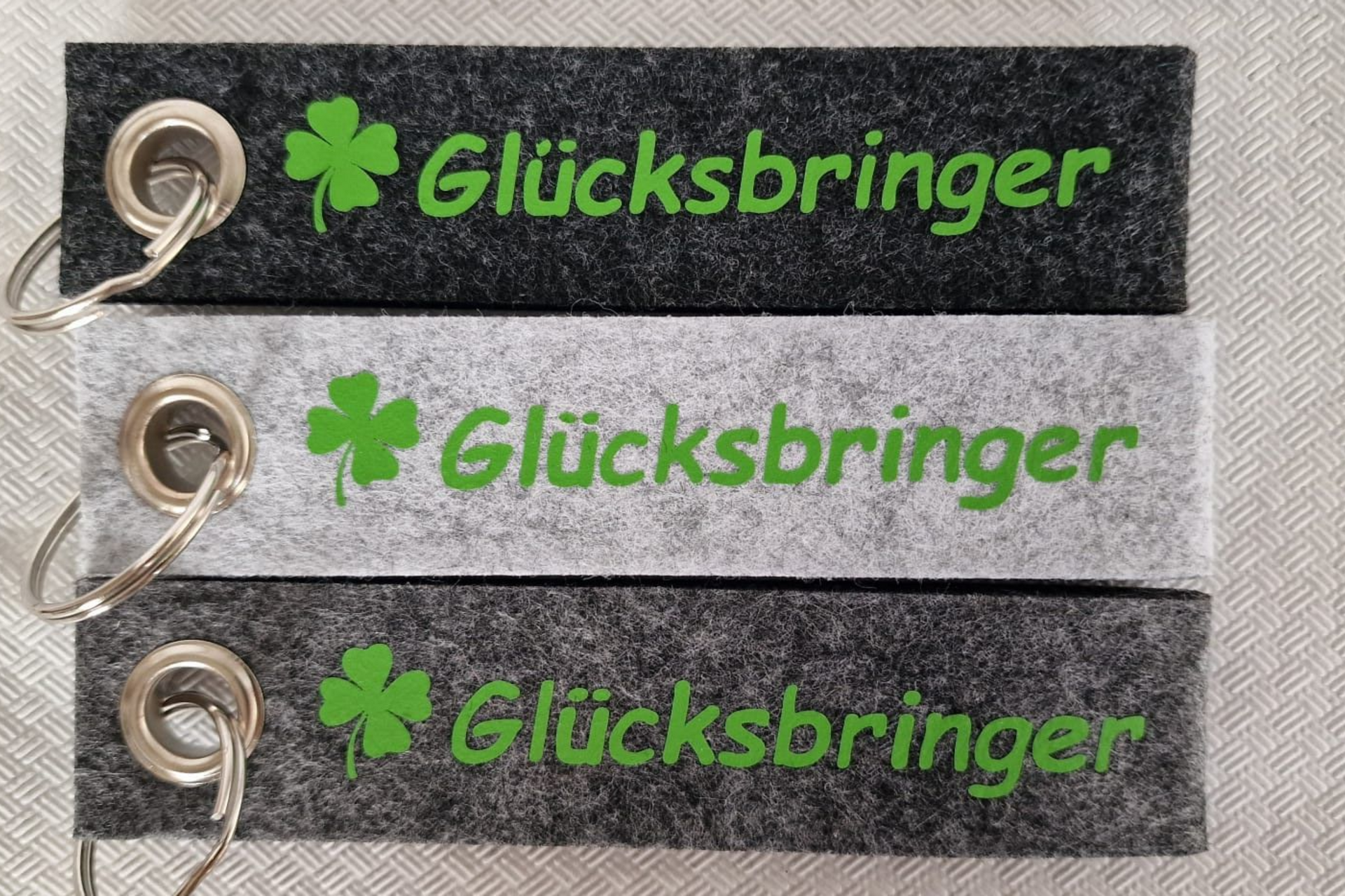 Drei Filz-Schlüsselanhänger, grau und schwarz, mit grünem „Glücksbringer“ und Kleeblatt.