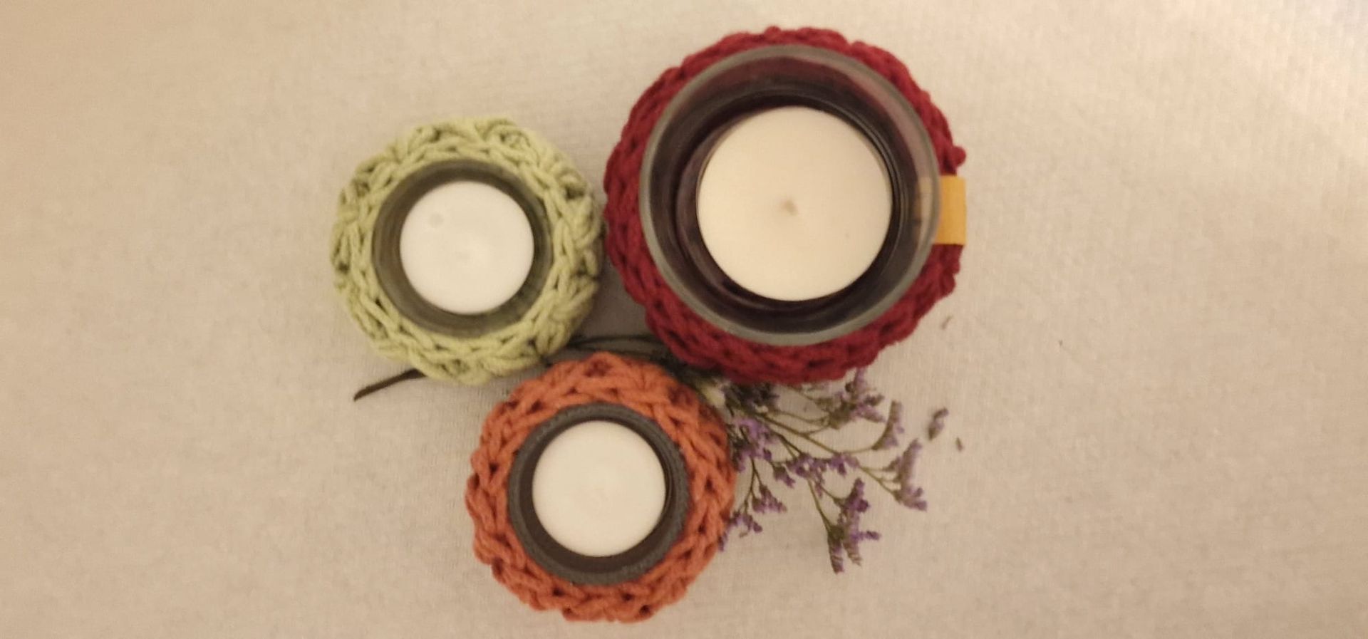 Drei gehäkelte Kerzenhalter in Grün, Rot und Orange, jeweils mit einer brennenden weißen Kerze auf einer weißen Oberfläche mit Lavendel.
