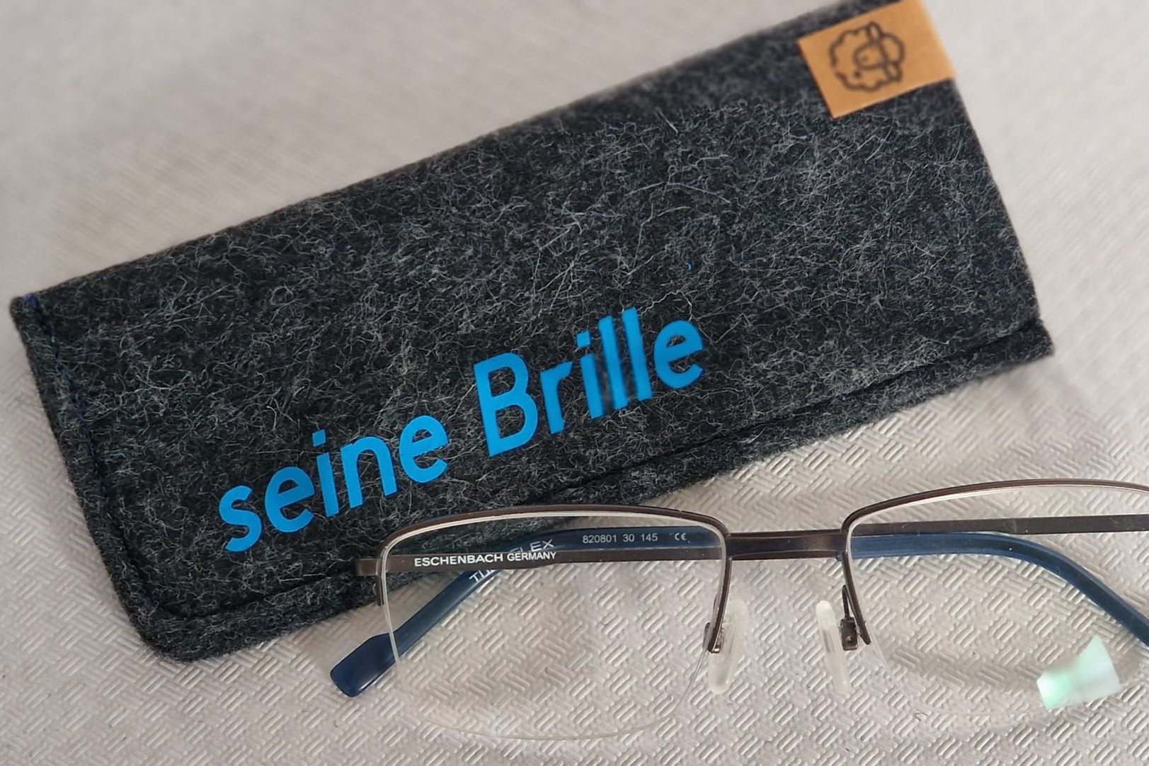 Graues Brillenetui mit der Aufschrift „seine Brille“ in Blau, daneben eine Brille.