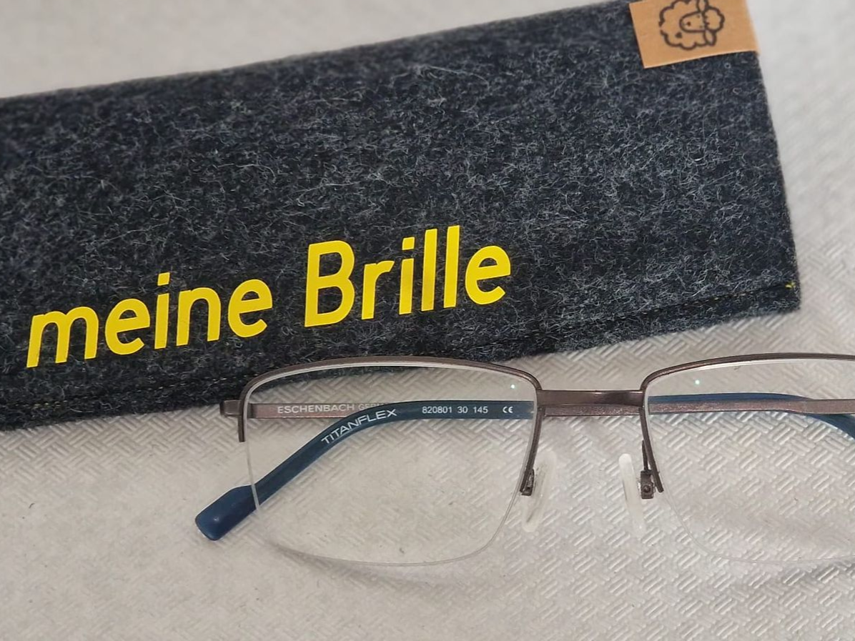 Brille und Etui. Graues Etui mit der Aufschrift „meine Brille“ in Gelb, Brille mit blauen Bügeln auf weißem Untergrund.