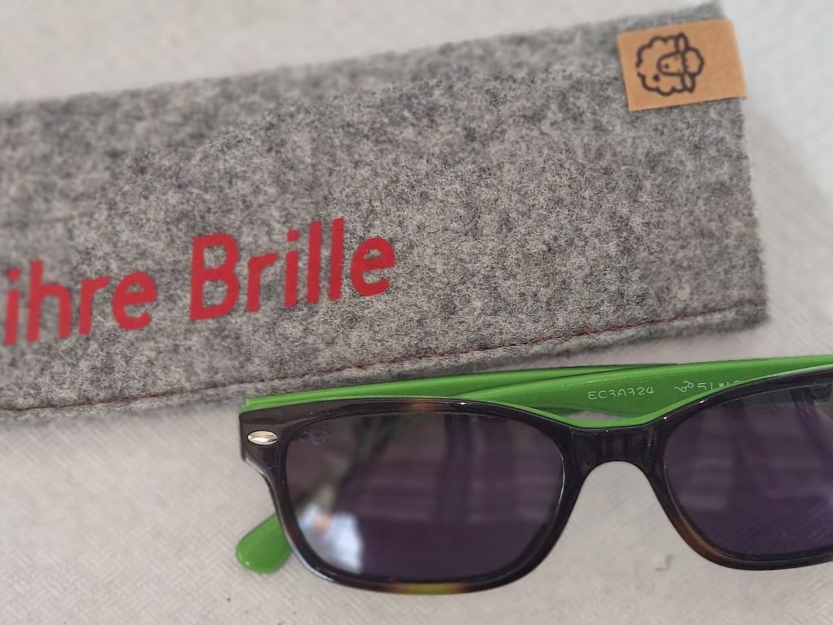 Sonnenbrille mit grünen Bügeln, daneben ein graues Brillenetui aus Filz mit der Aufschrift „Ihre Brille“.