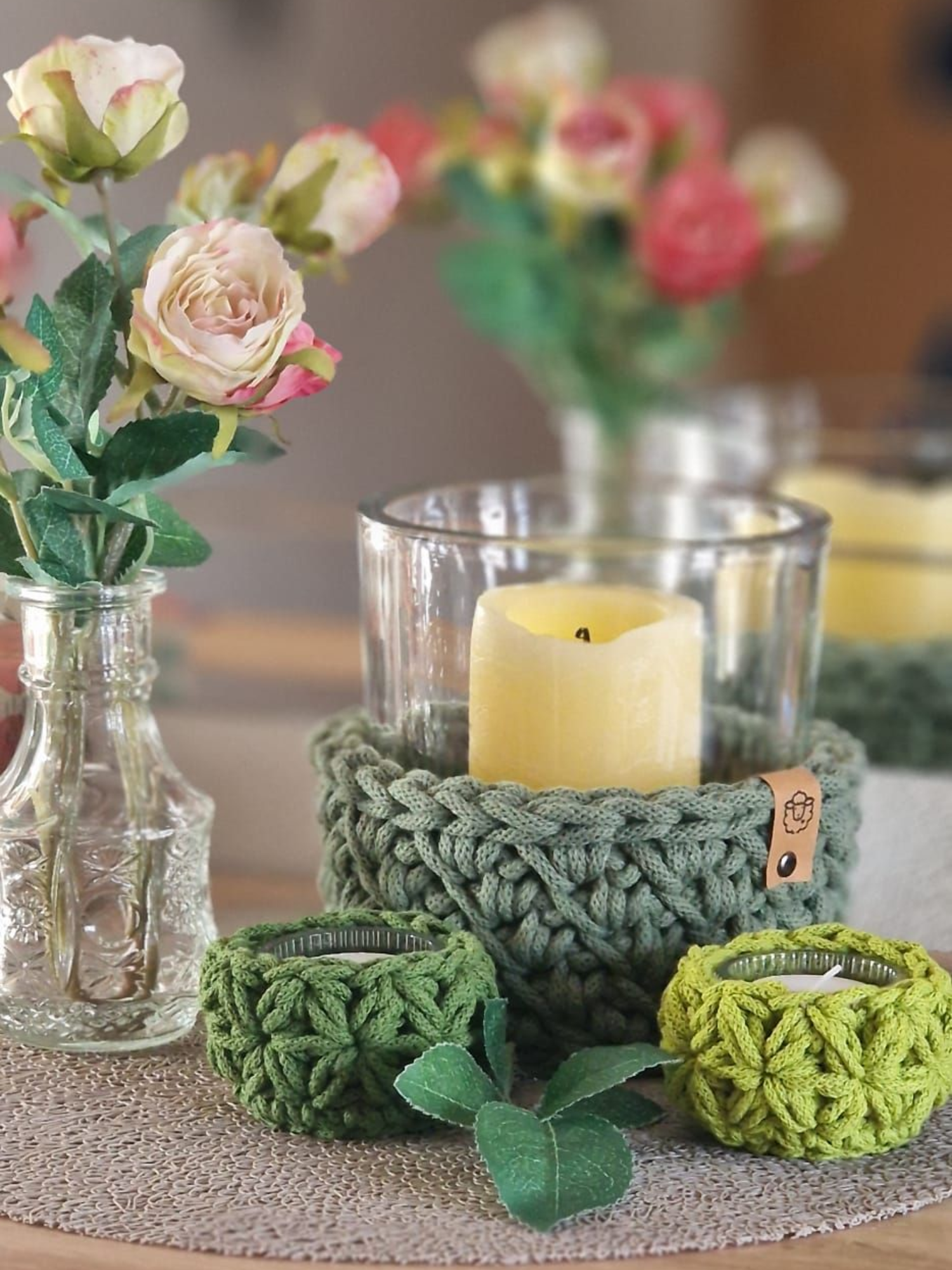 Gehäkelte grüne Kerzenhalter mit Kerzen, Blumen in Vasen auf einem Tisch.