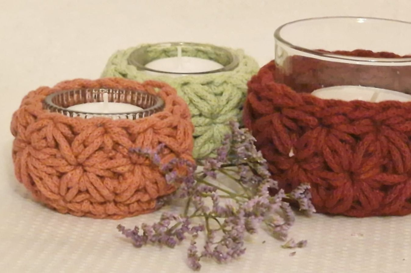 Drei gehäkelte Kerzenhalter in Orange, Grün und Rot, in denen jeweils eine brennende Kerze steht.