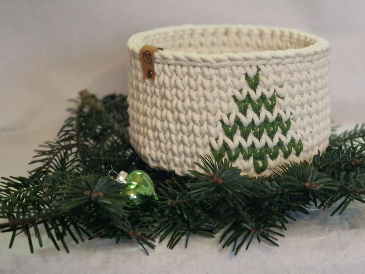 Gehäkelter, cremefarbener Korb mit aufgesticktem grünem Weihnachtsbaum, umgeben von immergrünen Zweigen.