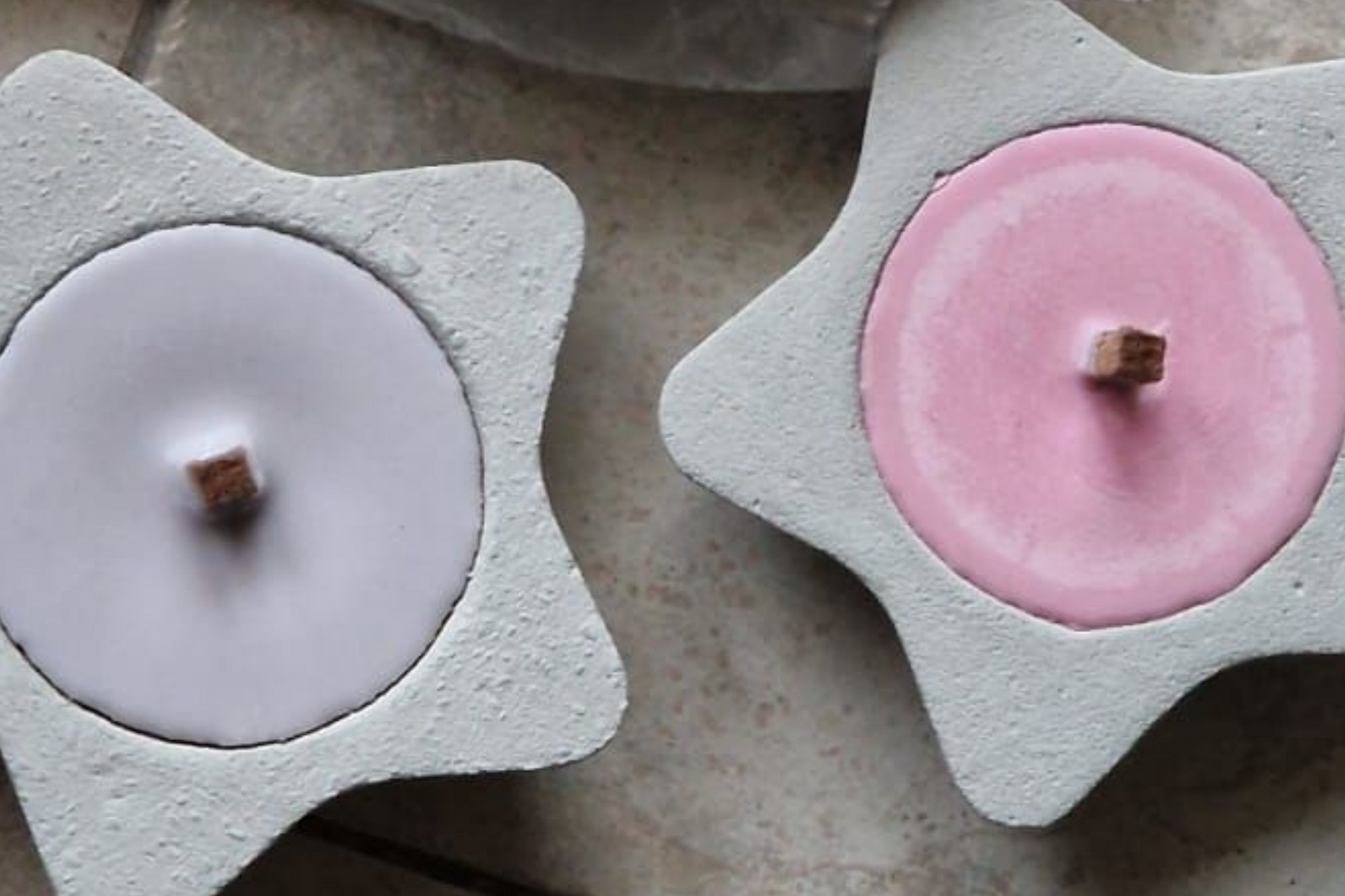Zwei sternförmige Kerzenhalter, einer rosa, einer lavendelfarben, mit hölzernen Dochthaltern in der Mitte.