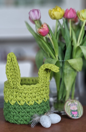 Eine kleine Vase mit Blumen in einem gehäkelten Korb auf einer gehäkelten Unterlage; braune und beige Farbtöne.