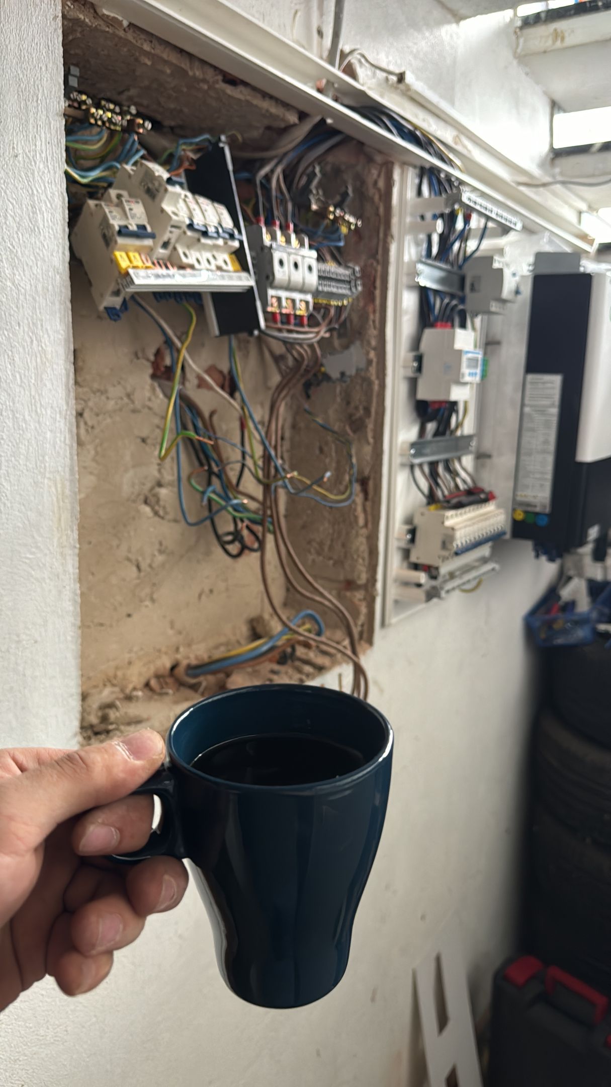 Eine Hand hält einen schwarzen Becher vor freiliegenden elektrischen Leitungen und Schalttafeln an einer Wand.