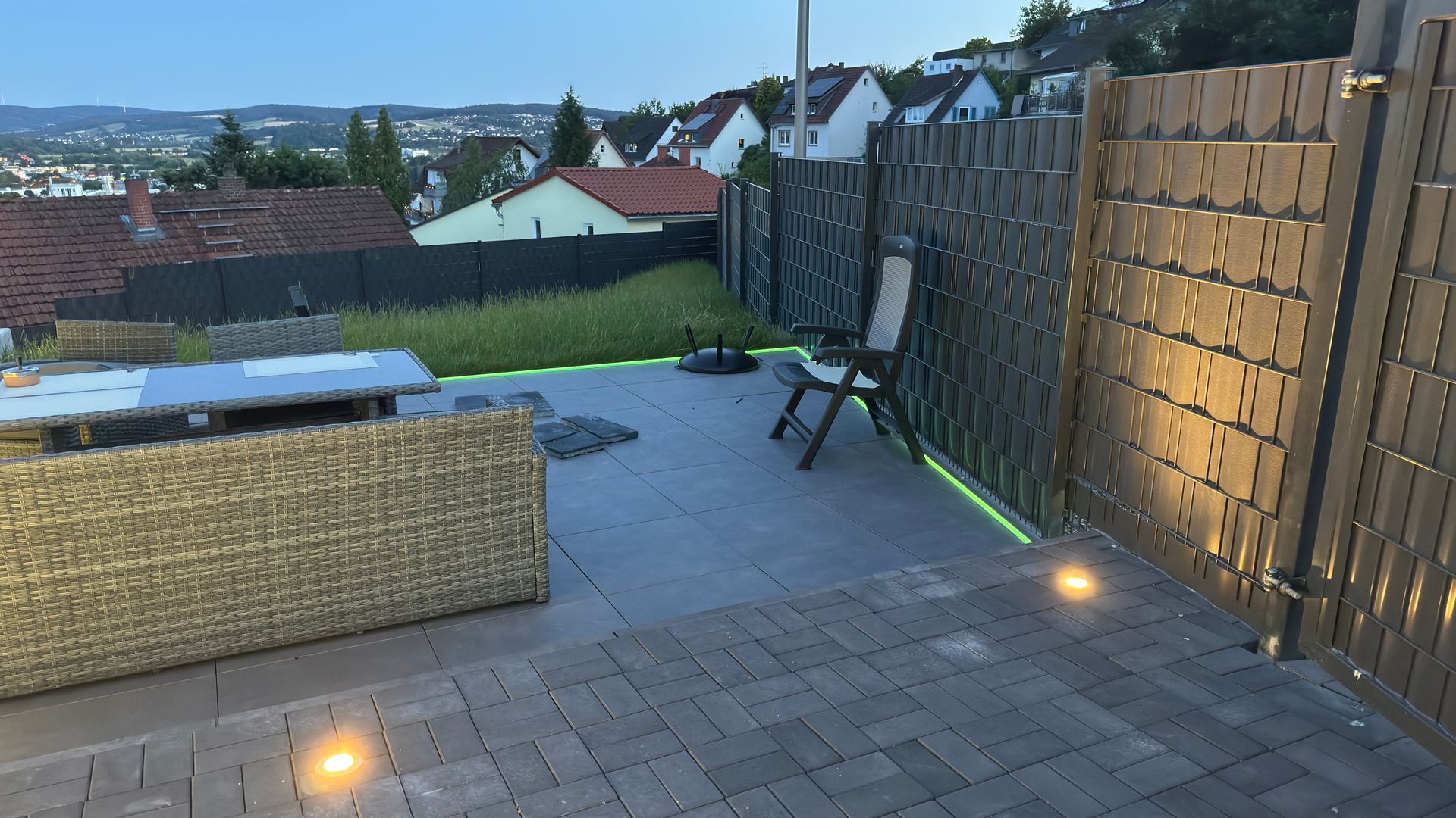 Hinterhof in der Abenddämmerung mit Außenbeleuchtung, geflochtener Bank, Holzzaun und fernen Häusern hinter einem grasbewachsenen Hang