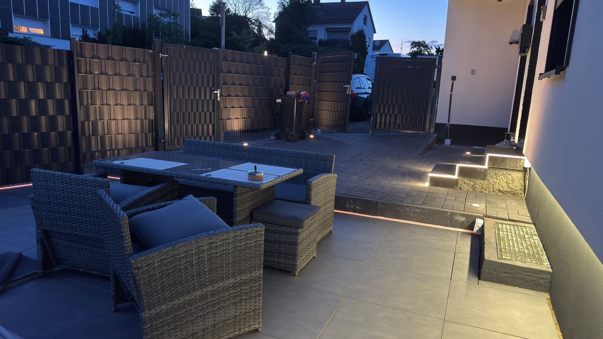 Terrasse im Hinterhof in der Abenddämmerung mit Korbmöbeln, einem Tisch, einem Zaun und beleuchteten Stufen neben einem Haus.
