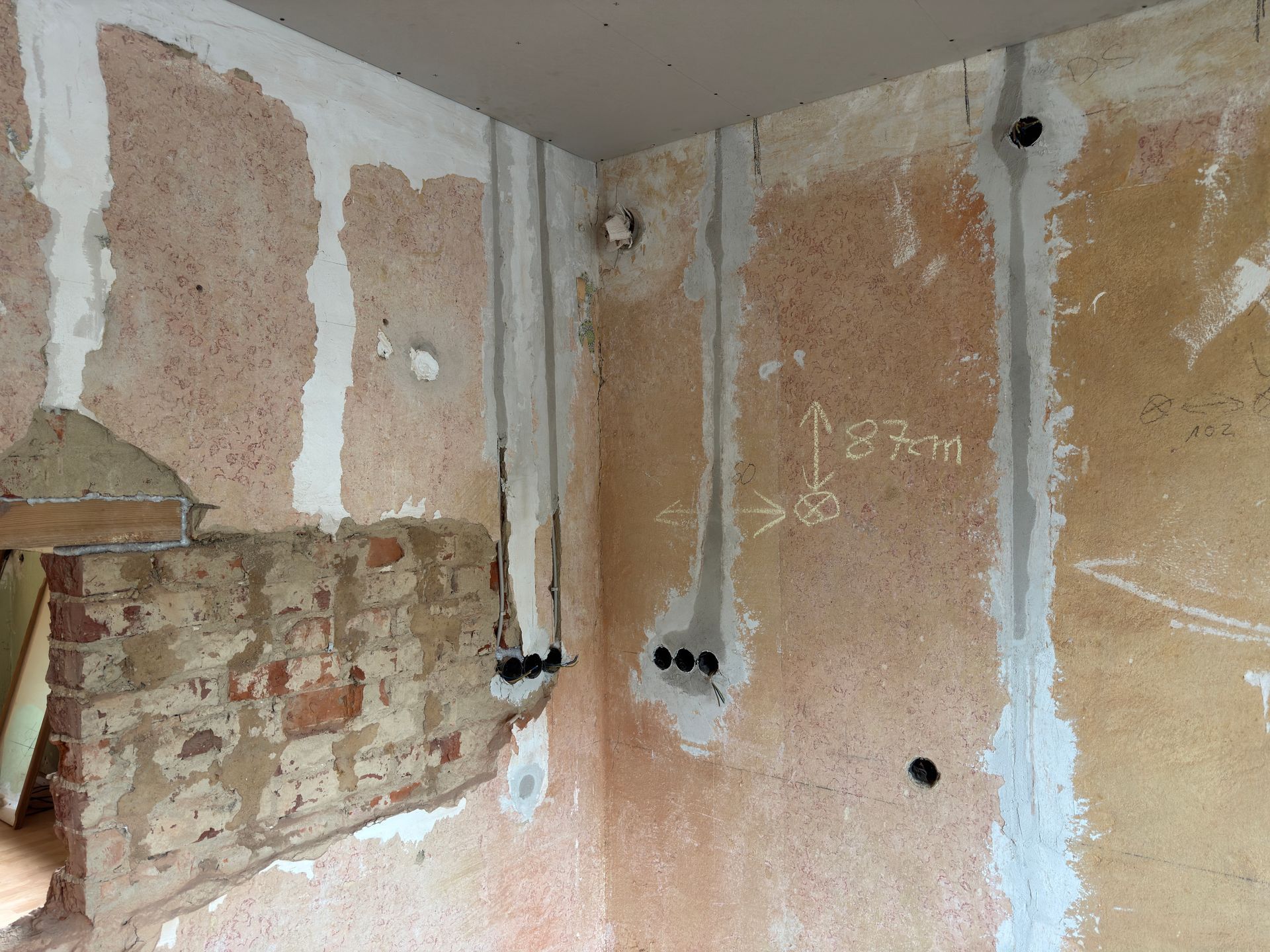 Teilweise freigelegte Gipskartonwände in einem Zimmer, mit freiliegendem Putz, Kabeldurchführungen und fleckigen beigen Wänden.