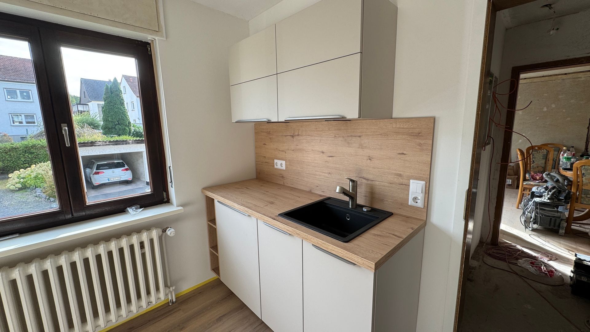 Kleine Küche mit weißen Schränken, schwarzem Spülbecken, beigefarbenem Fliesenspiegel und einem Fenster über einem Heizkörper.