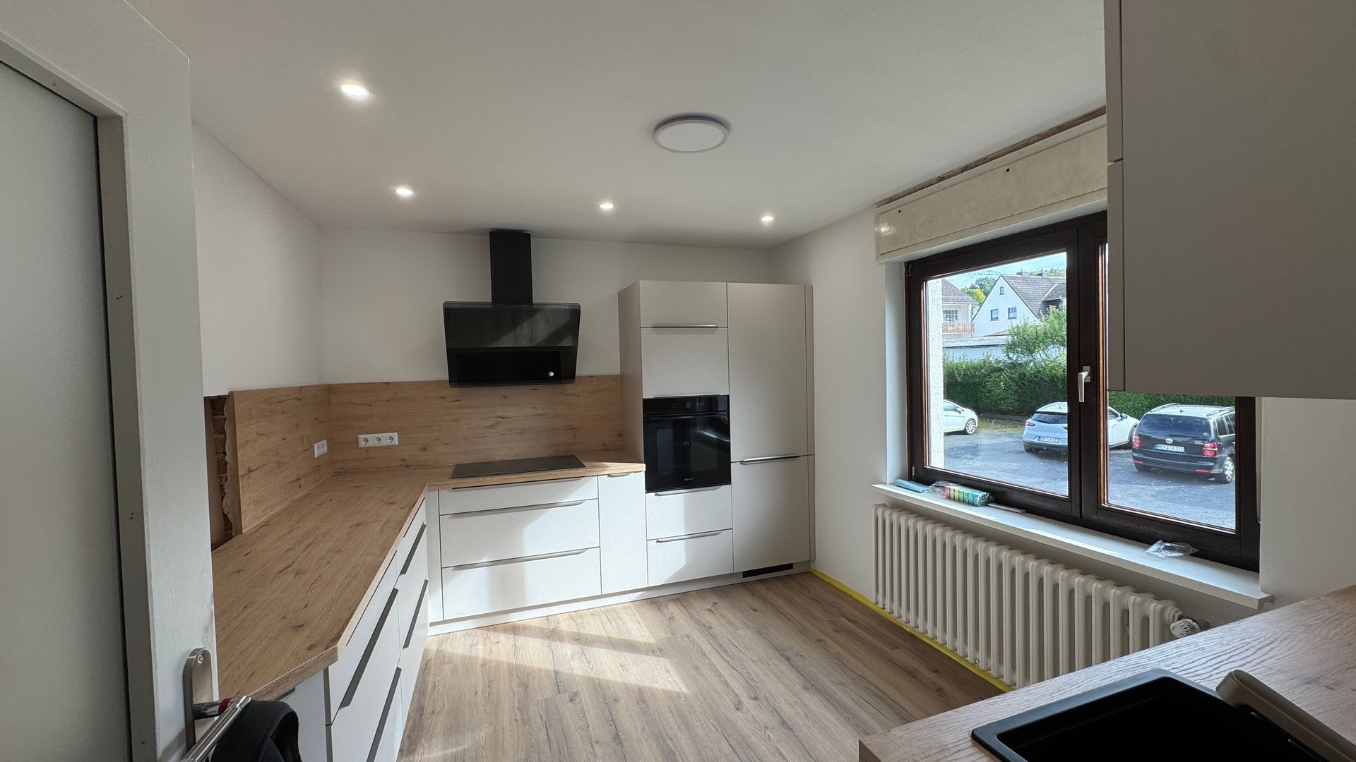 Helle, moderne Küche mit weißen Schränken, Holzboden, schwarzen Elektrogeräten und einem großen Fenster.