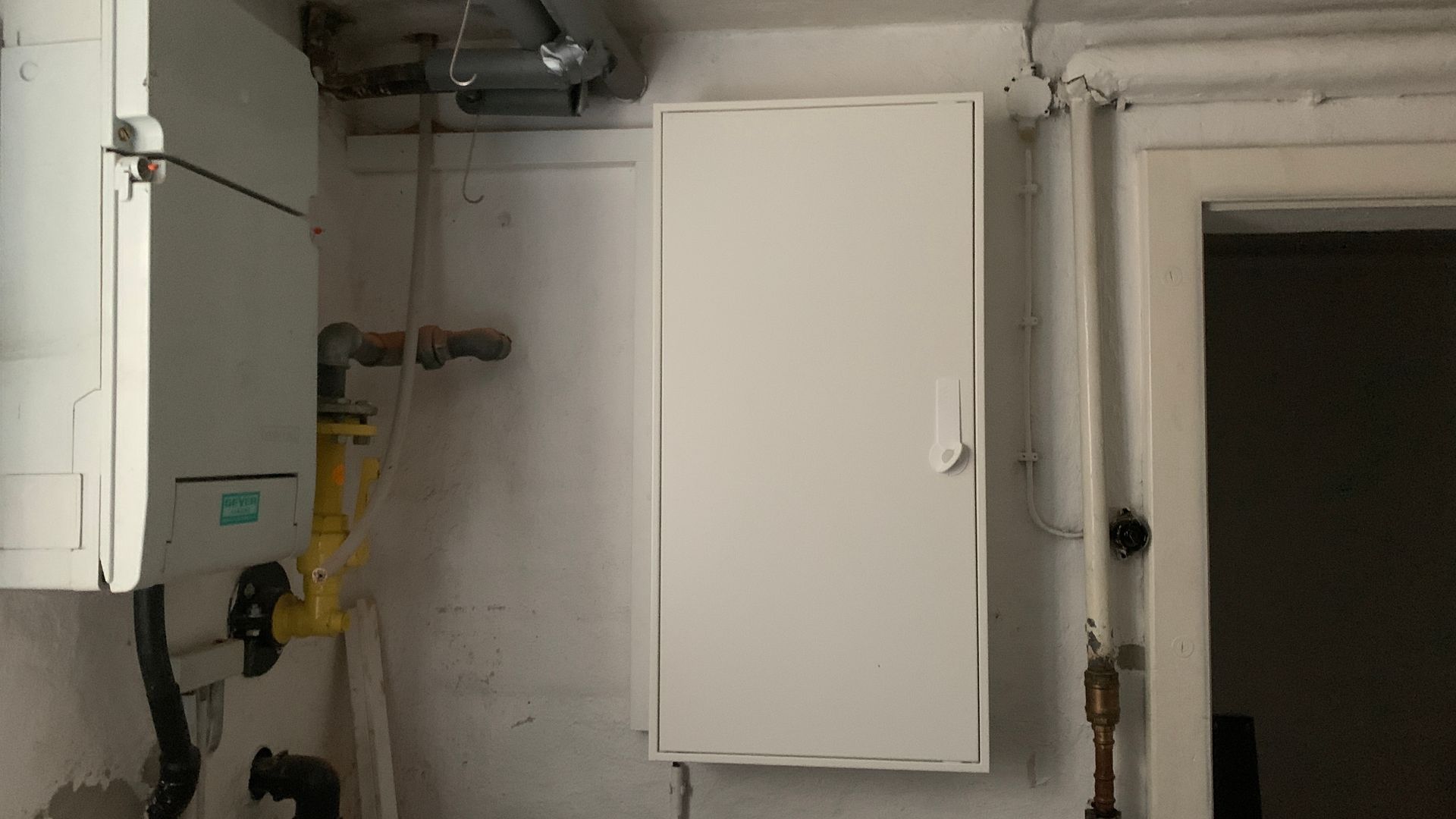 Hauswirtschaftsraum mit weißen Wandschränken, Rohren und einer offenen Tür.