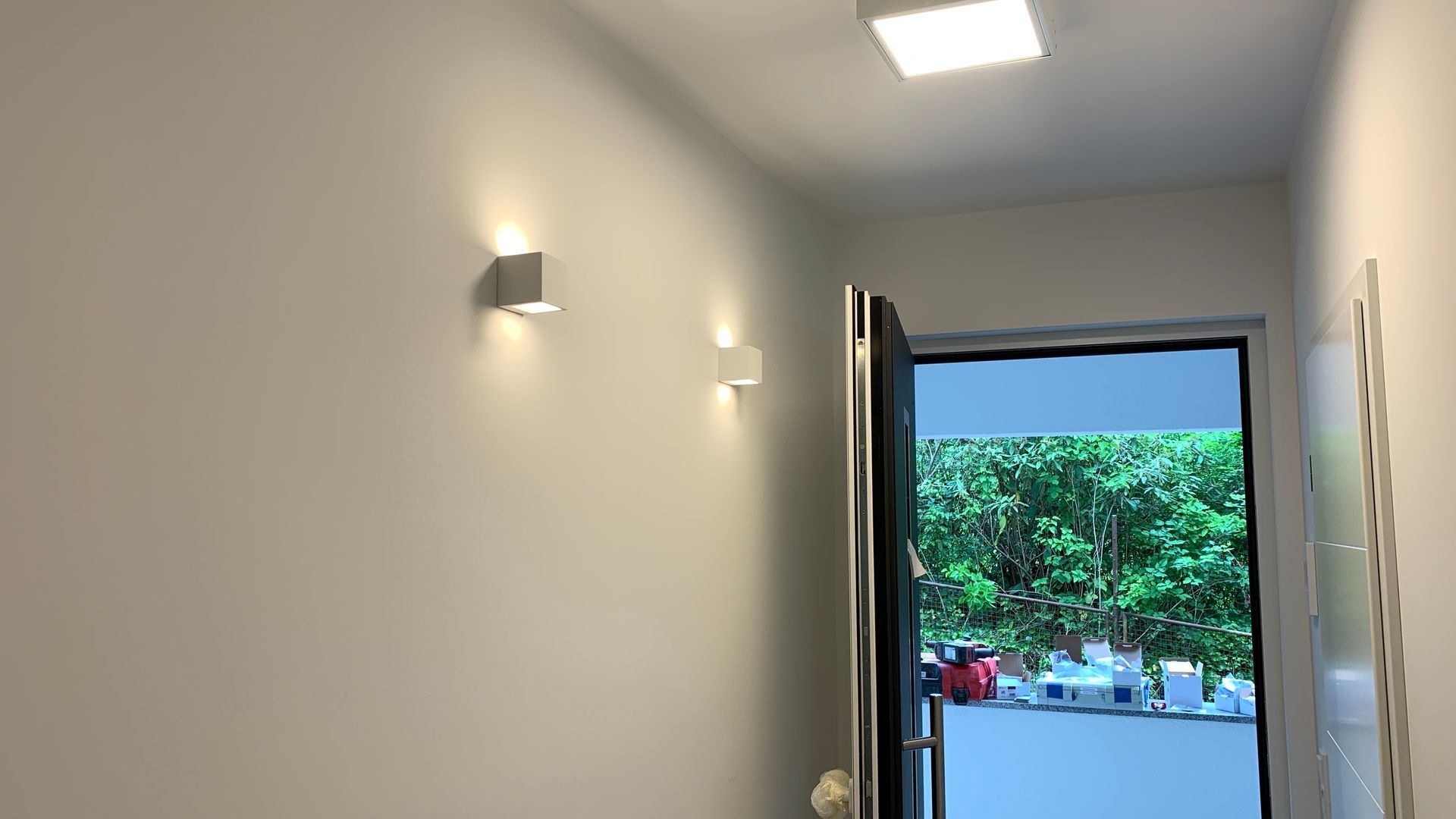 Ein heller Flur mit Wandleuchten führt zu einer offenen Tür mit Blick auf die Bäume draußen.