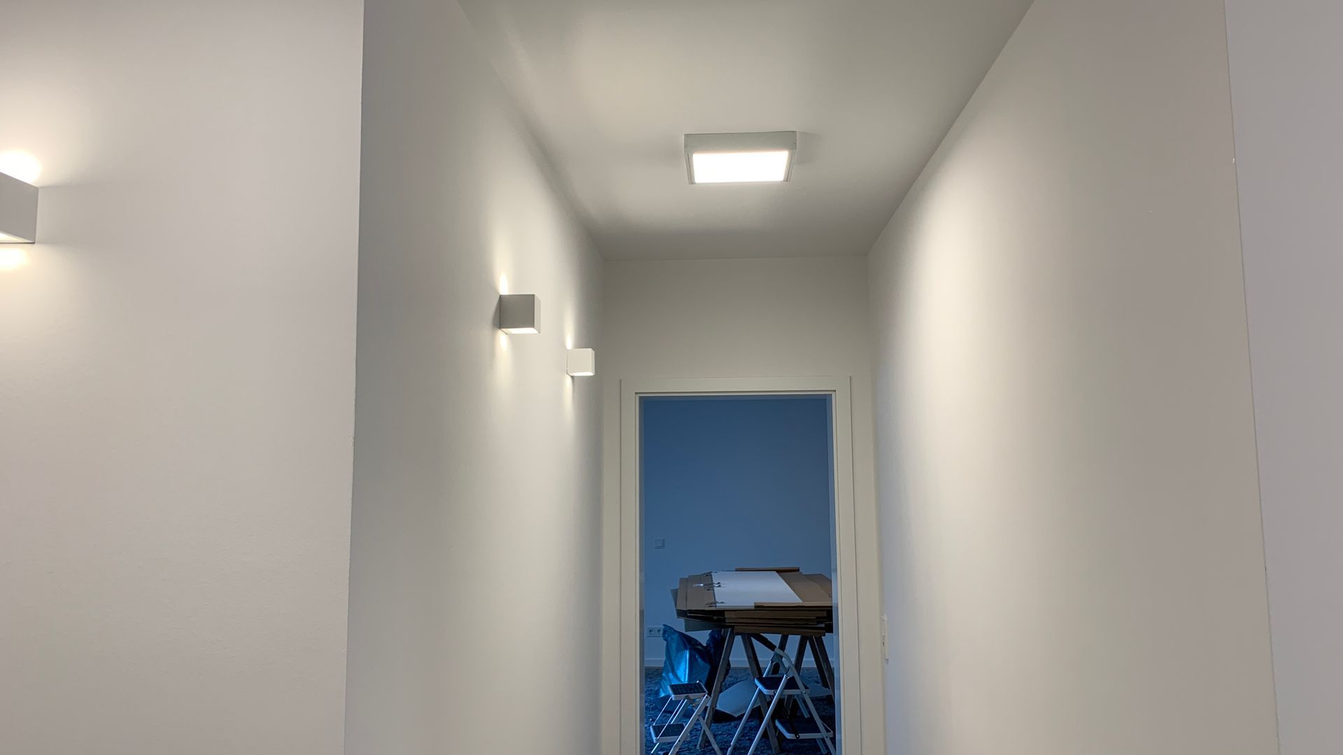 Ein schmaler, weißer Flur mit Deckenleuchte führt zu einem blau gestrichenen Raum mit Tisch und Stühlen.