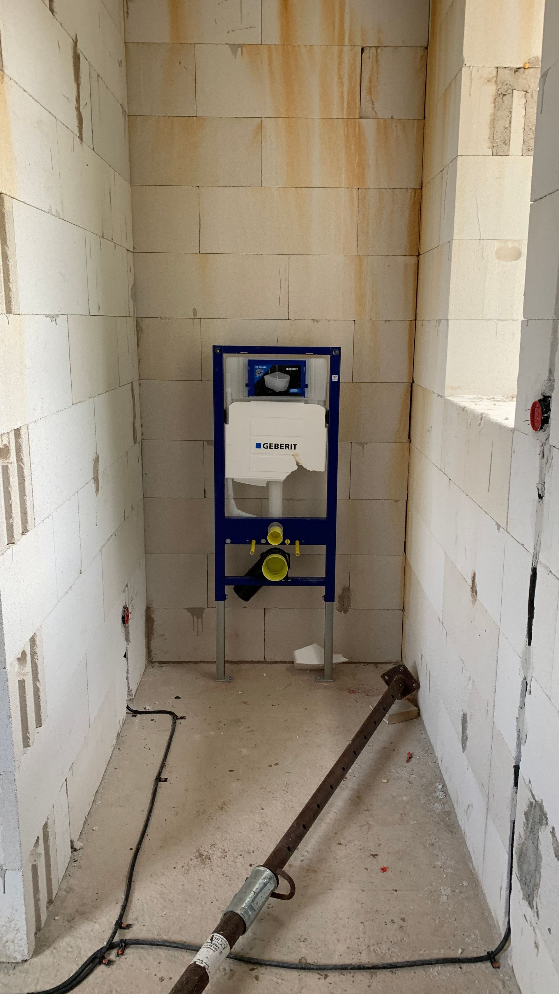 Ein blauer Toilettenspülkasten wurde in einem schmalen, unfertigen Badezimmer mit freiliegenden Mauersteinwänden und Bauschutt auf dem Boden installiert.