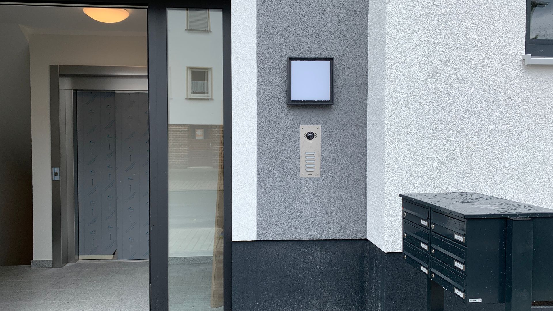 Wohnungseingang mit Gegensprechanlage, Wandleuchte und Briefkästen neben der Tür.