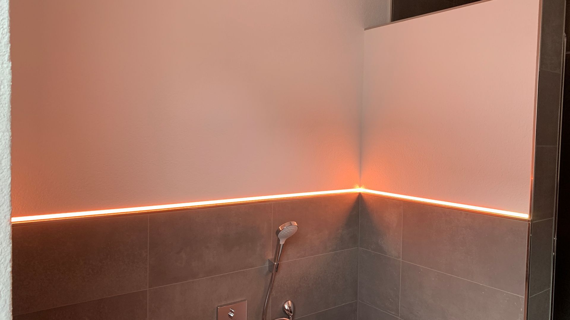 Badezimmerecke mit grau gefliester Wand, Wandnische und warmweißer LED-Beleuchtung entlang des Spritzschutzes.