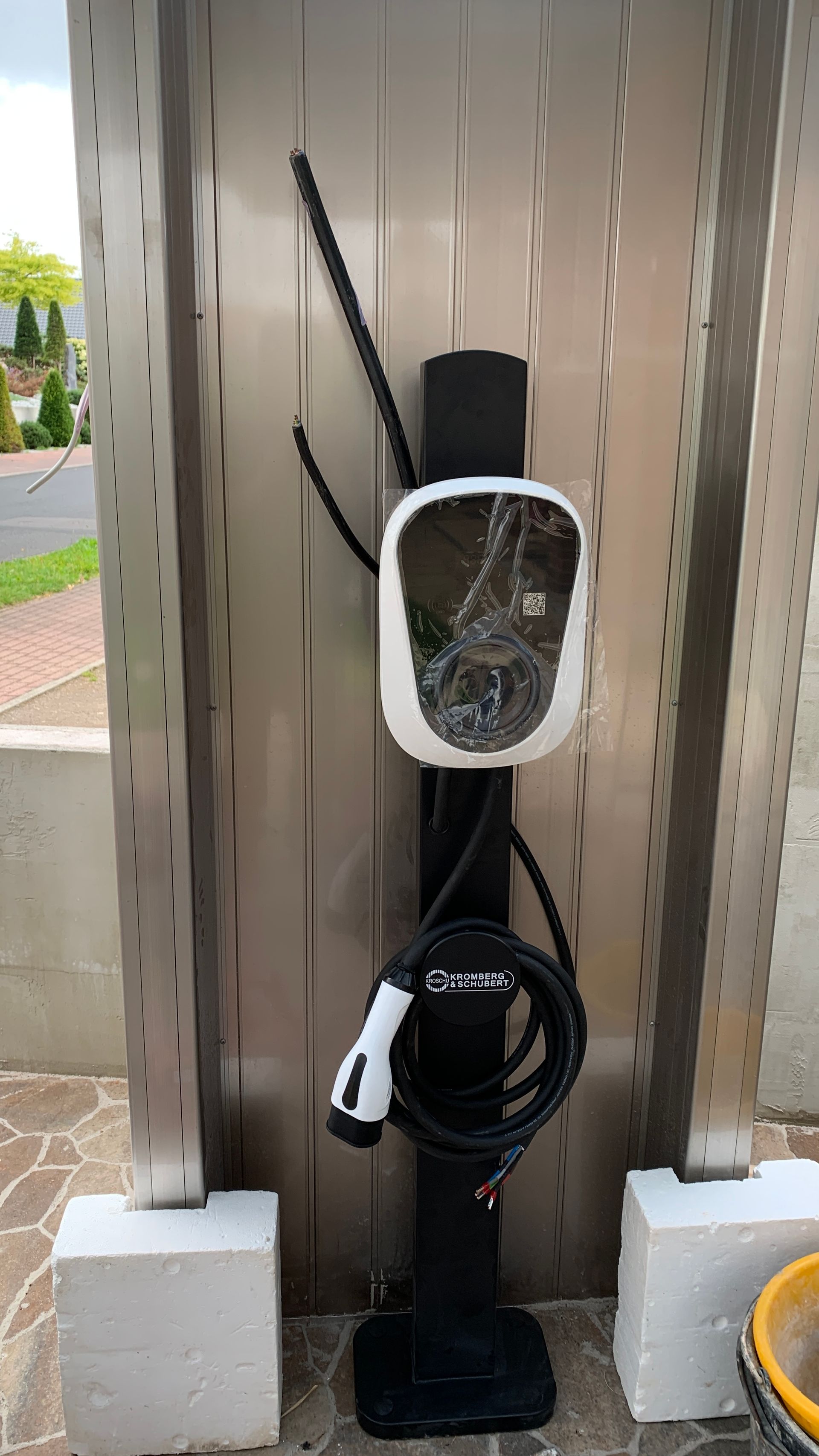 An einem dunklen Pfosten ist eine Wandladestation für Elektrofahrzeuge angebracht, mit aufgerolltem Kabel und seitlichen Antennen neben einem Parkplatz.