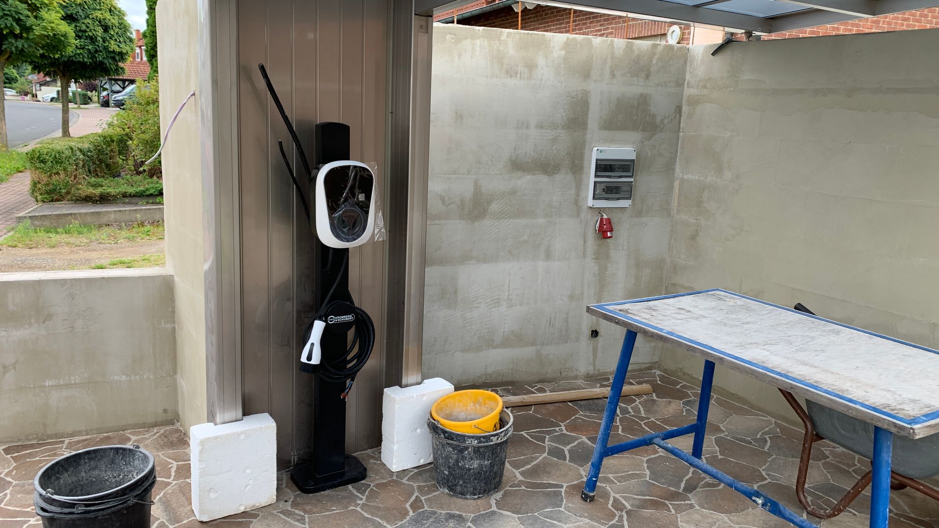 Außennutzungsbereich mit einem schwarzen Wasserspender, Eimern, einem Tisch und einem an der Wand montierten Zählerkasten.