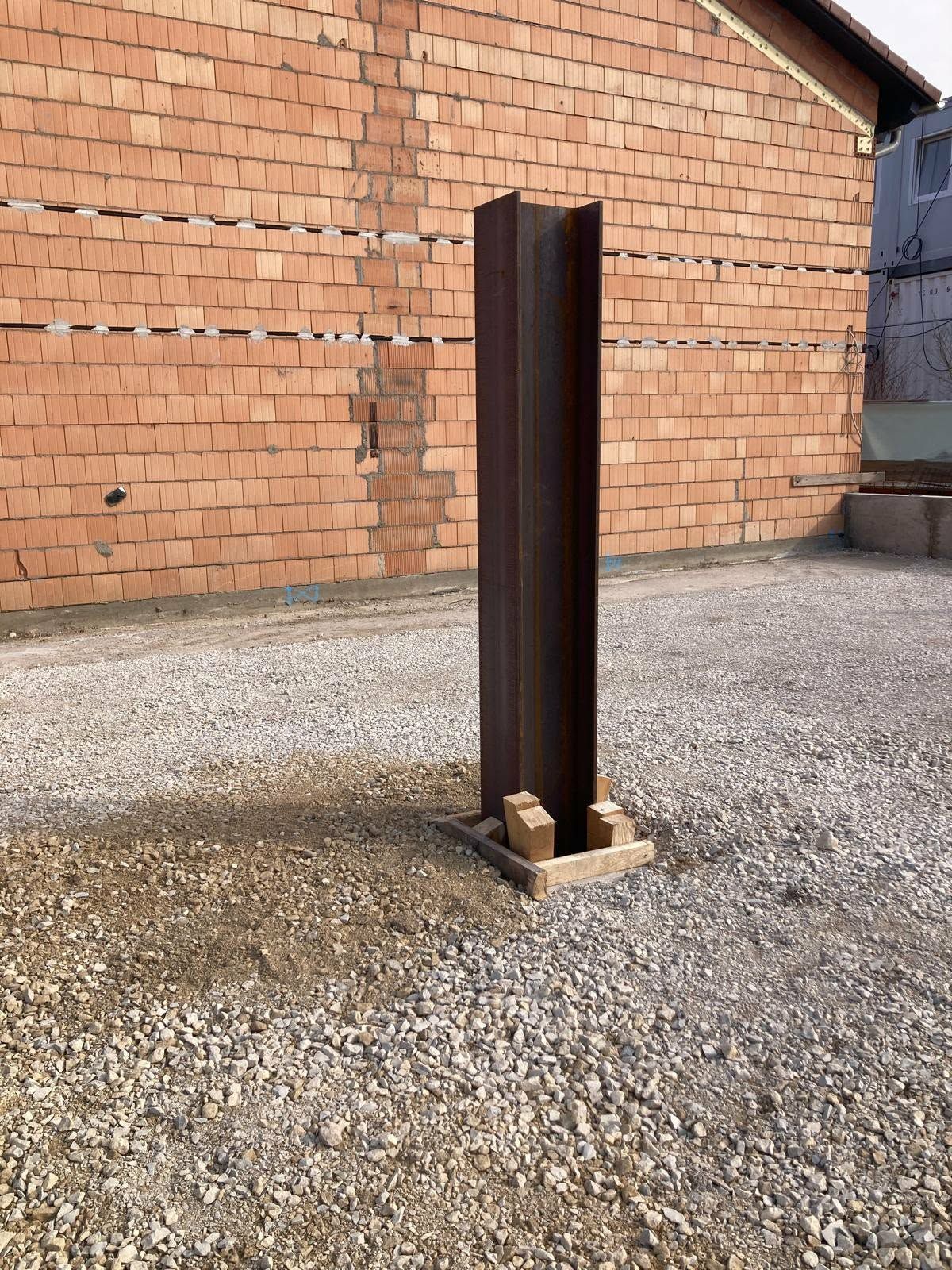 Ein hoher, dunkelbrauner Stahlträger in H-Form steht aufrecht auf einem quadratischen Betonsockel in einem Kieshof vor einer Backsteinmauer.