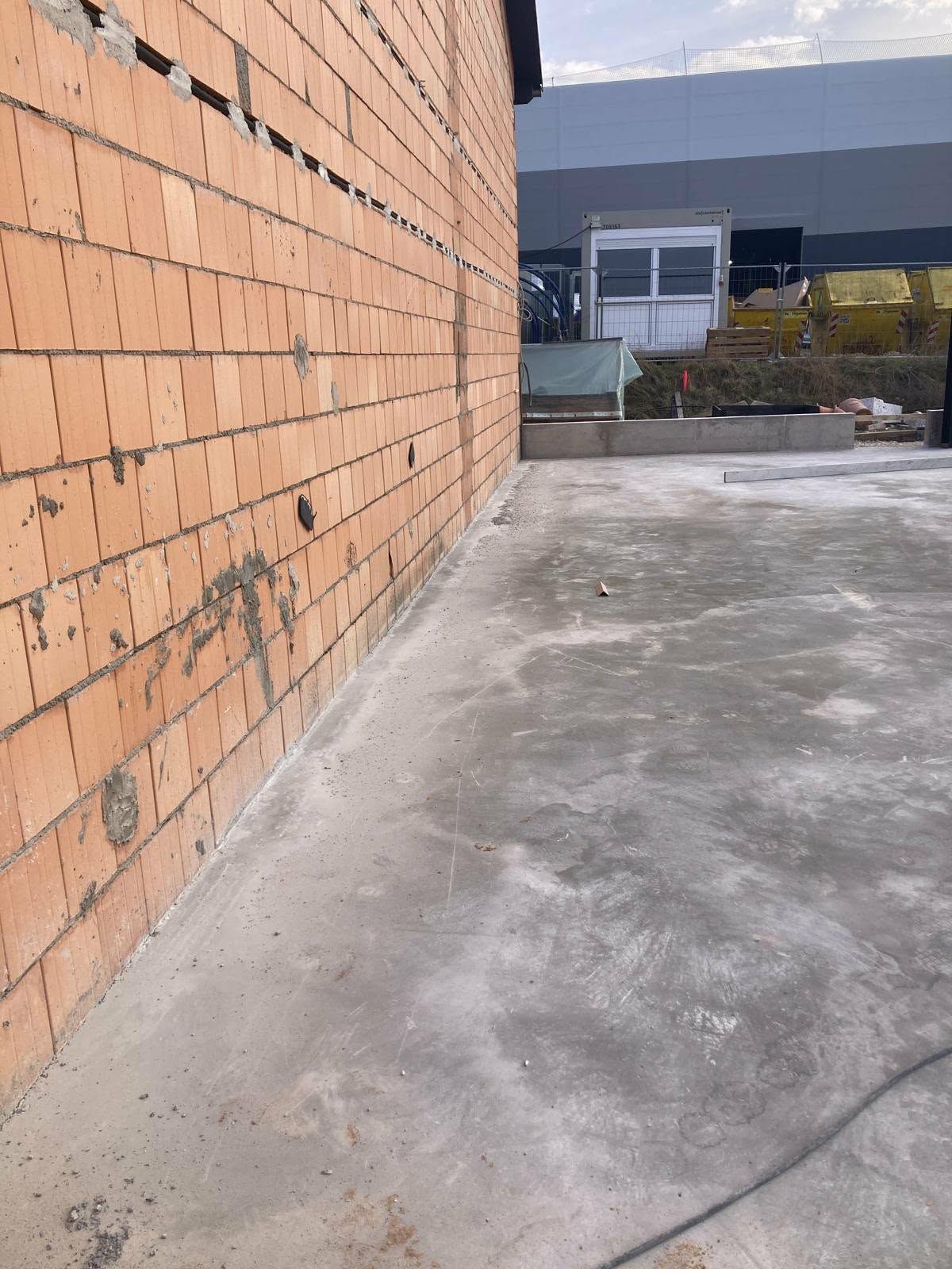 Eine Ziegelmauer verläuft entlang einer Betonplatte auf einer Baustelle; im Hintergrund ist ein entferntes Gebäude zu sehen.