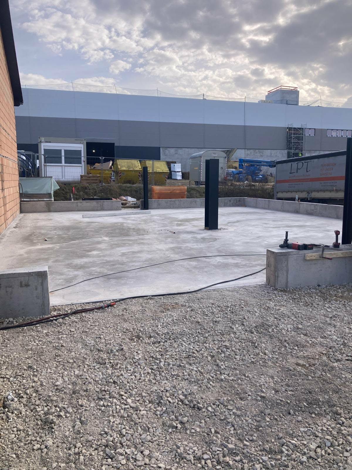 Baustelle mit einer Betonfundamentplatte und Stahlstützsäulen vor einem großen Industriegebäude.