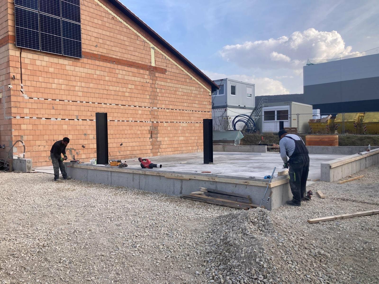 Zwei Arbeiter bereiten auf einer Baustelle ein Betonfundament vor einer gemauerten Gebäudewand vor.