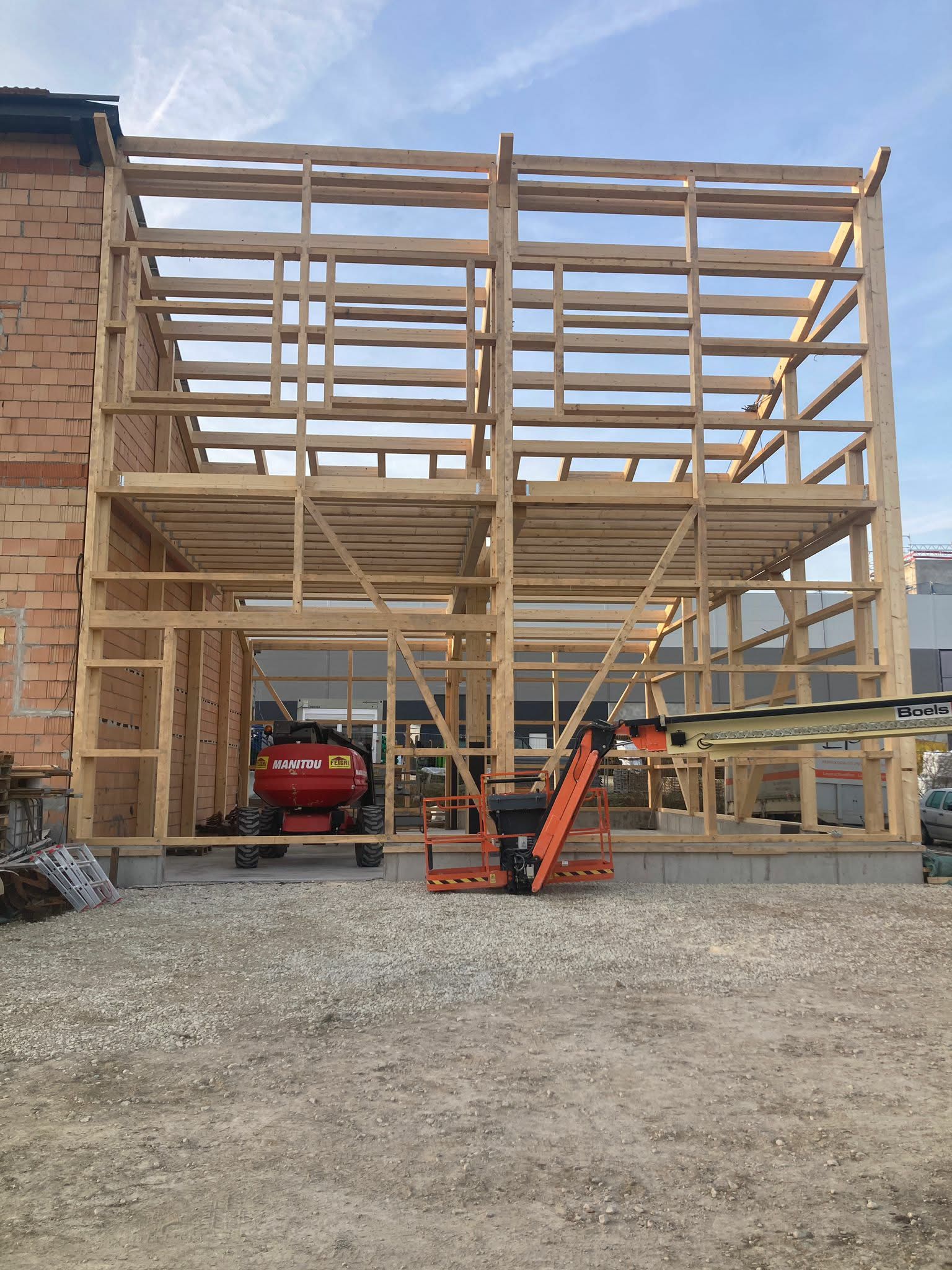 Neben einem Backsteingebäude befindet sich eine im Bau befindliche Holzrahmenkonstruktion, in deren Inneren ein orangefarbener Bauaufzug parkt.