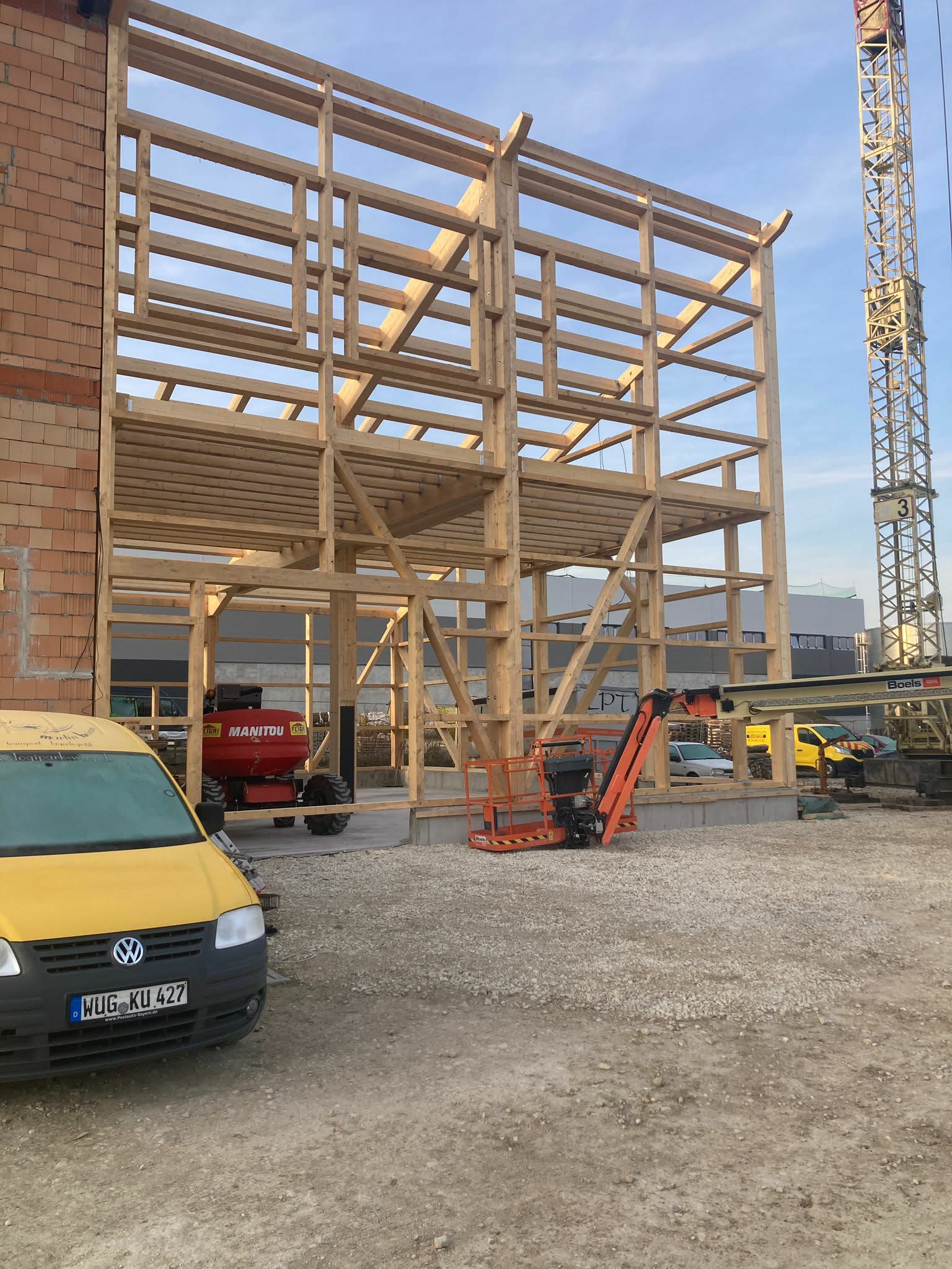 Ein im Bau befindliches Fachwerkgebäude neben einer Backsteinmauer, auf dem ein gelber Lieferwagen und Baumaschinen zu sehen sind.