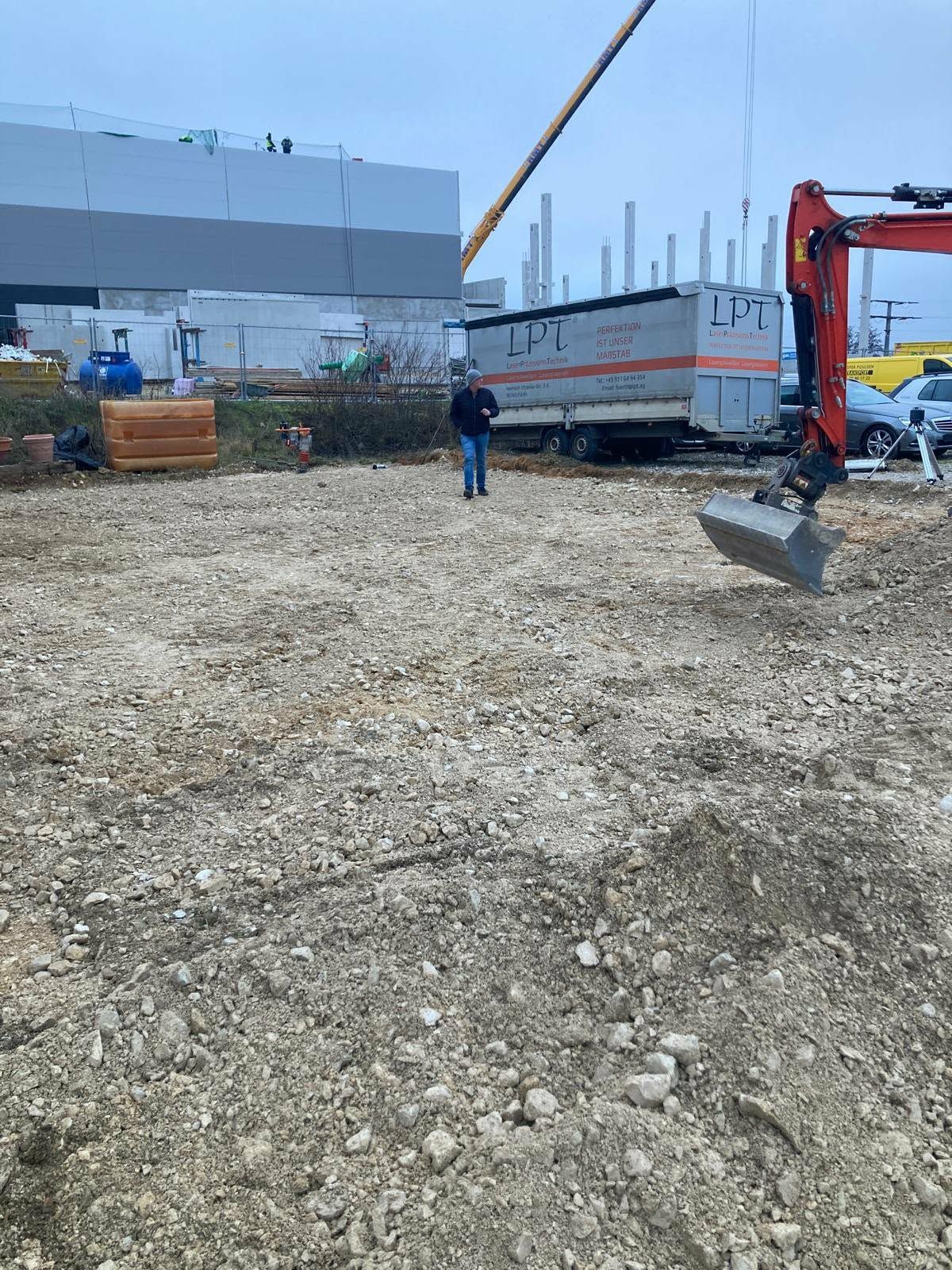 Eine Person steht auf einem Schotterplatz in der Nähe einer Baustelle mit einem Kran, einem Bagger und einem geparkten Anhänger.