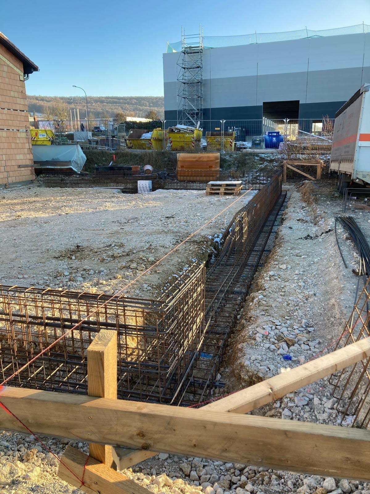 Bewehrungskörbe für Gebäudefundamente liegen auf einer Schotterbaustelle; im Hintergrund ist ein großes Industriegebäude zu sehen.