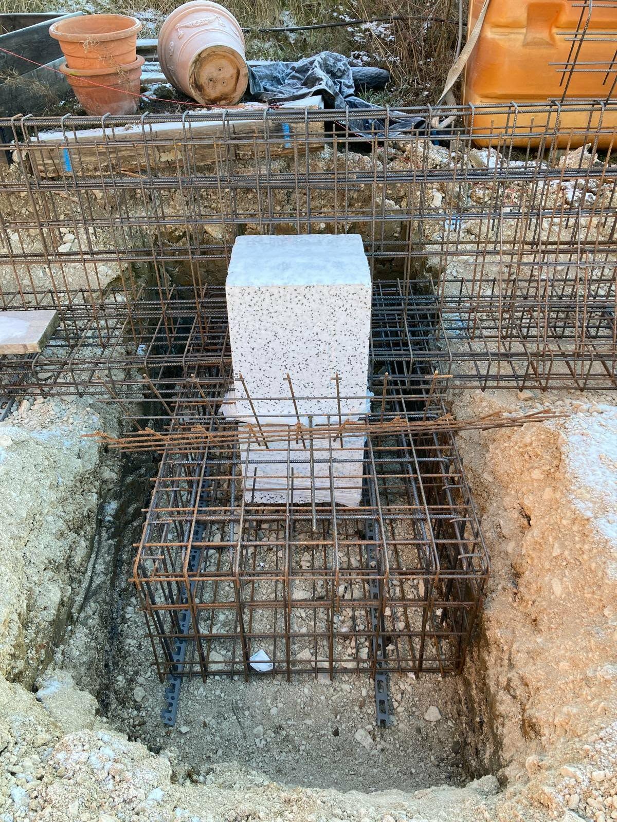 Ein quadratischer weißer Block steht auf einem Metallbewehrungsrahmen, der für ein Betonfundament auf einer unbefestigten Baustelle vorbereitet ist.