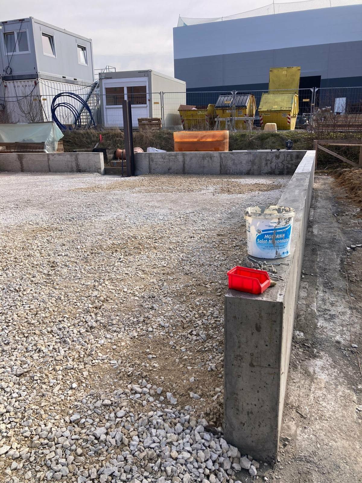 Eine mit Kies gefüllte Baustelle mit einer Betonfundamentmauer im Vordergrund, auf der ein Farbeimer und eine Farbwanne zu sehen sind.