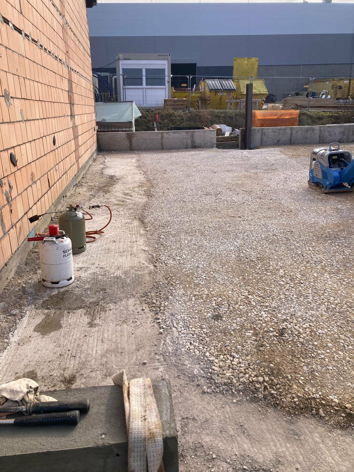 Eine mit Kies bedeckte Baustelle mit einer Backsteinmauer auf der linken Seite, einem blauen Stromgenerator und Geräten im Vordergrund.