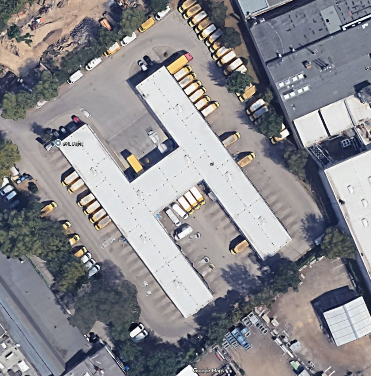 Luftaufnahme eines Parkplatzes mit mehreren gelben Bussen neben langen weißen Industriegebäuden.