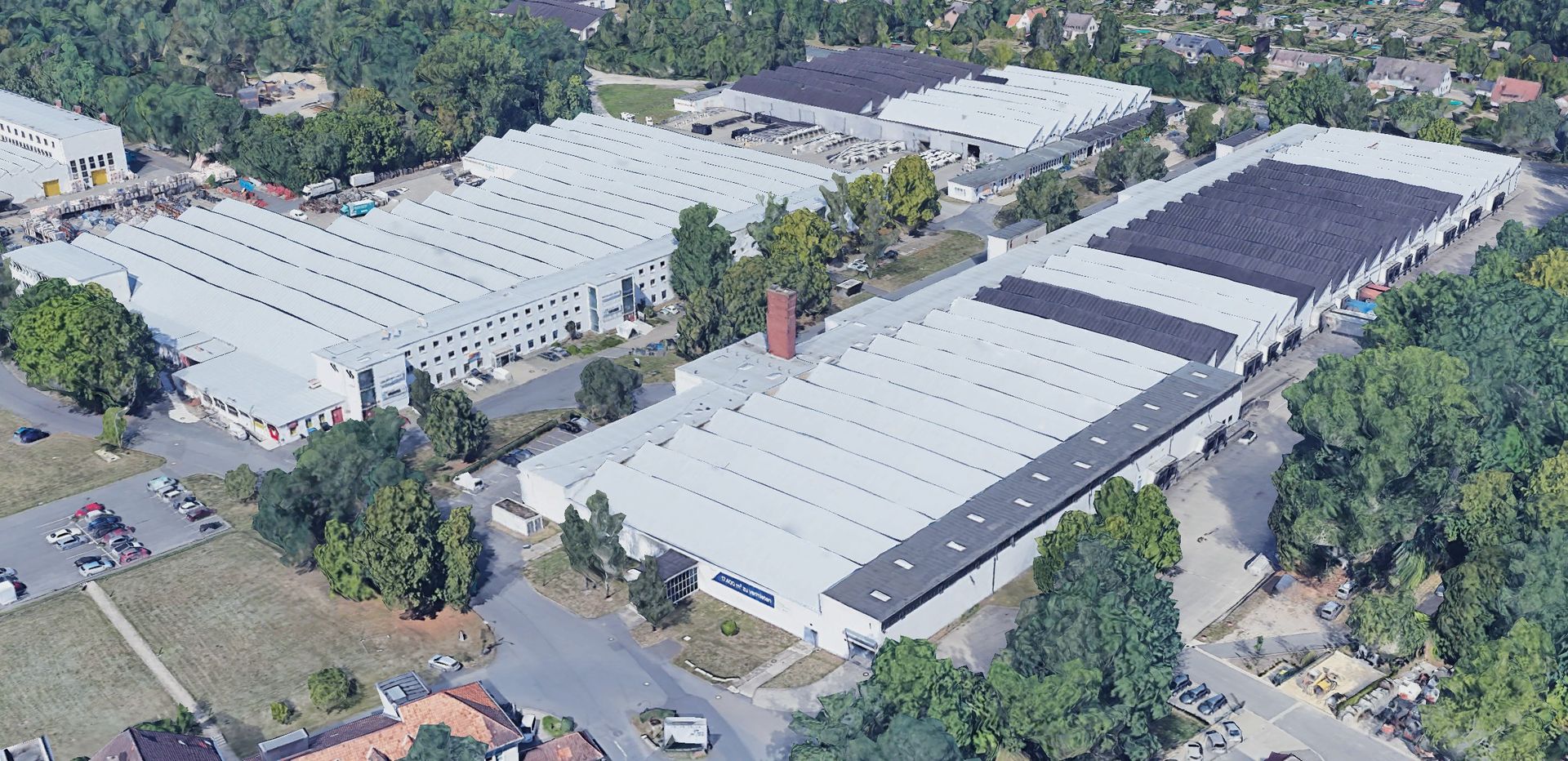 Luftaufnahme von großen Industriegebäuden mit weißen Dächern, umgeben von Bäumen und Straßen.