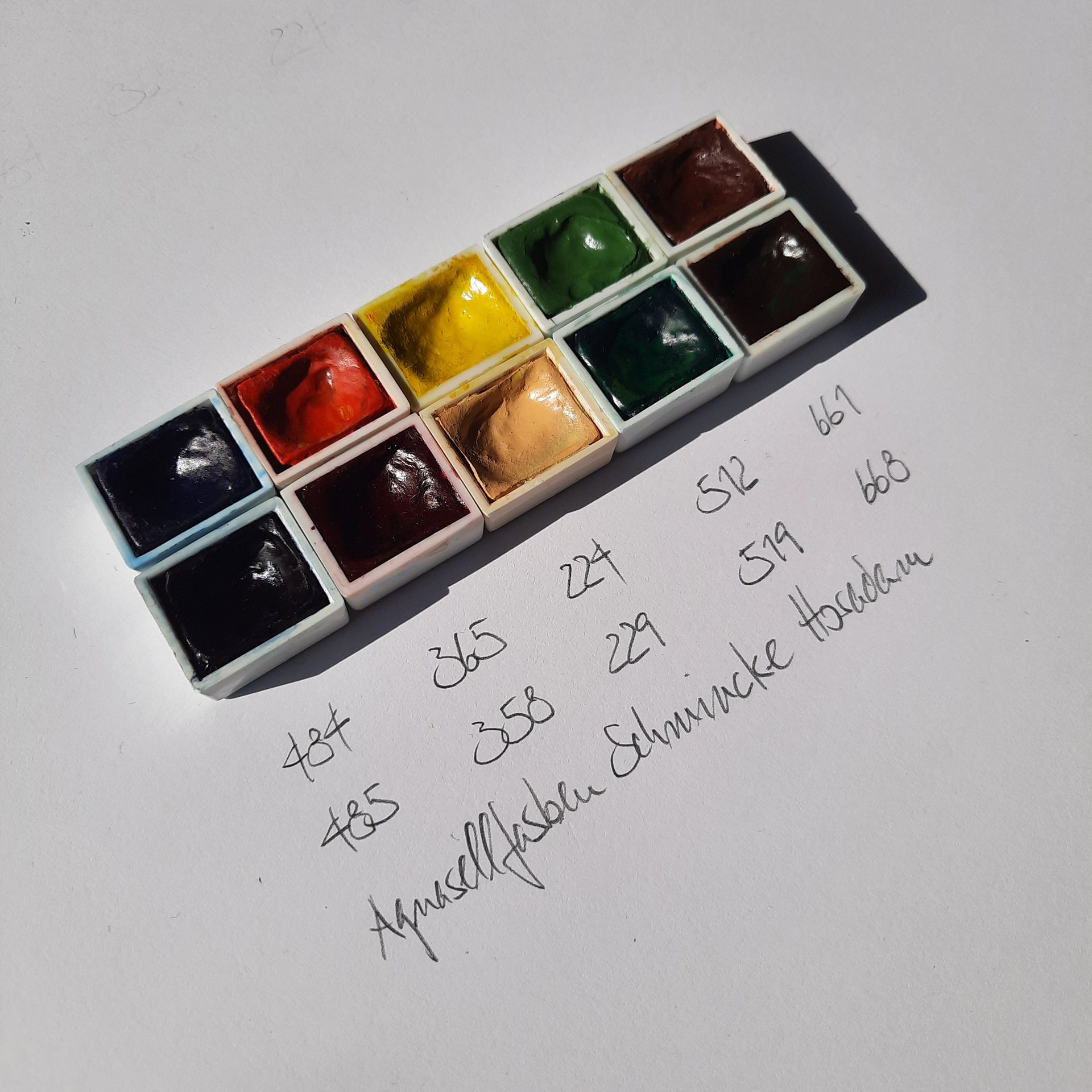 Zehn halbe Näpfchen Aquarellfarbe und davor von Hand geschrieben die Farbnummern der Marke Schmincke Horadam.