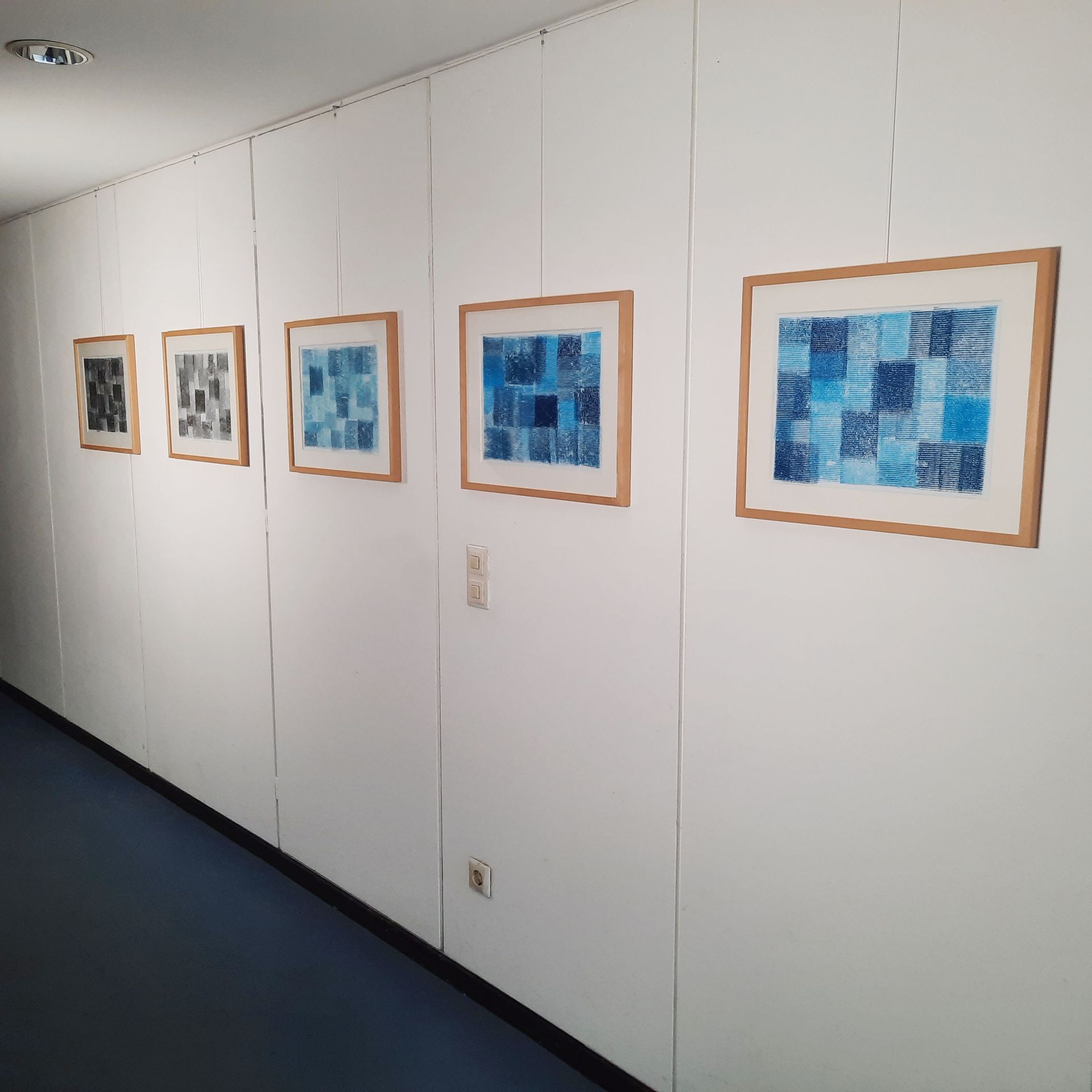 Wand eines Büroflurs mit einer Reihe von fünf Bildern im Querformat. Holzrahmen mit Passepartout und blauen /grauen Grafiken.
