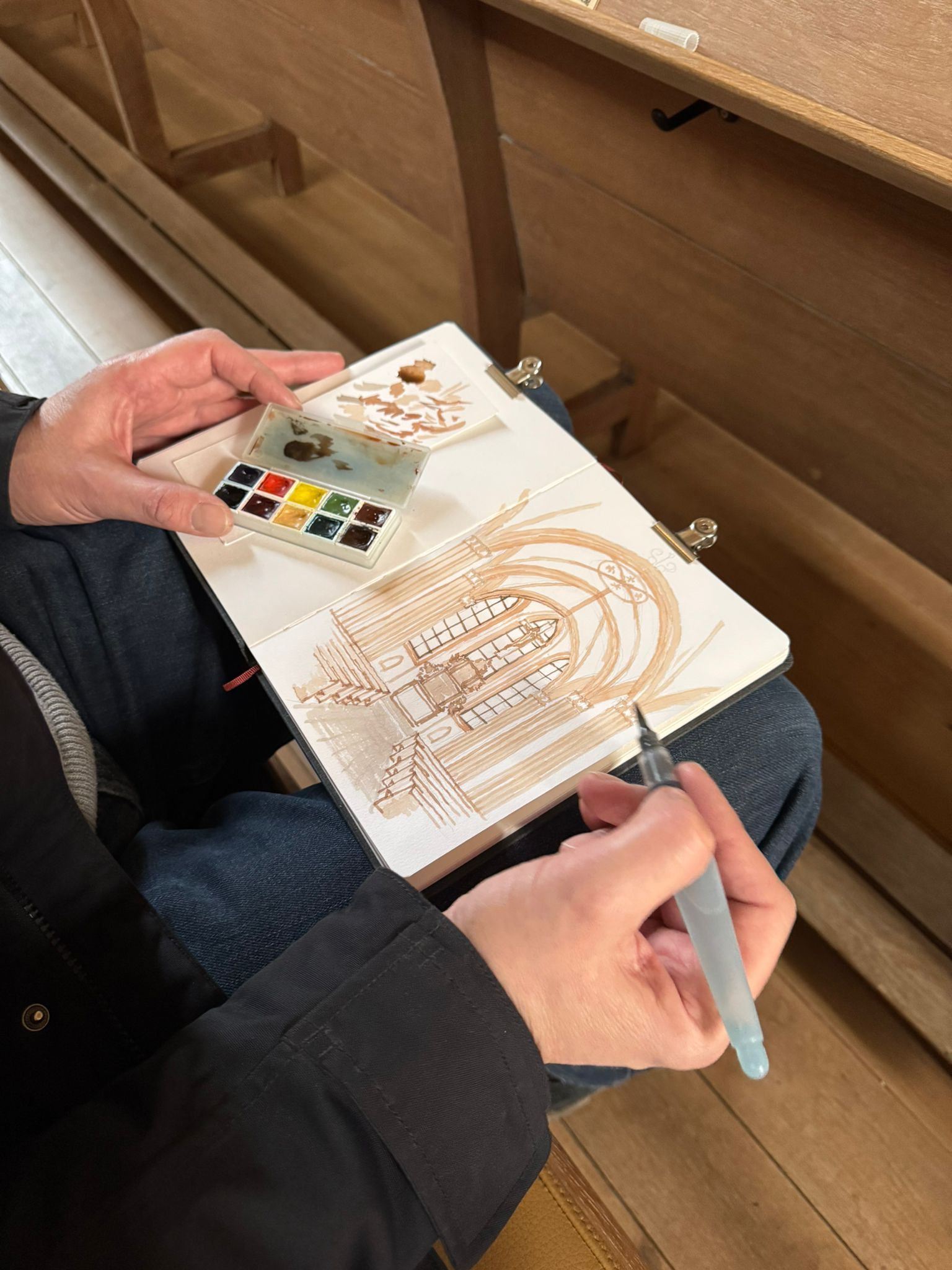 Man sieht mich auf einer Kirchenbank sitzend mit einem aufgeschlagenen Skizzenbuch auf den Knien und einer aquarellierten Zeichnung der Gewölbe des Kirchenraums. Am oberen Bildrand sieht man die schräge Ablage für Gesangbücher.