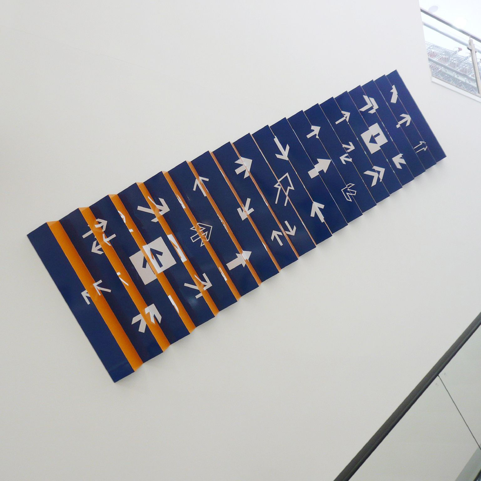 Plastisches Wandbild als Leporello neben einer Rolltreppe. Als Hintergrundfarben wechseln sich Blau und Orange ab, die Pfeile sind weiß.