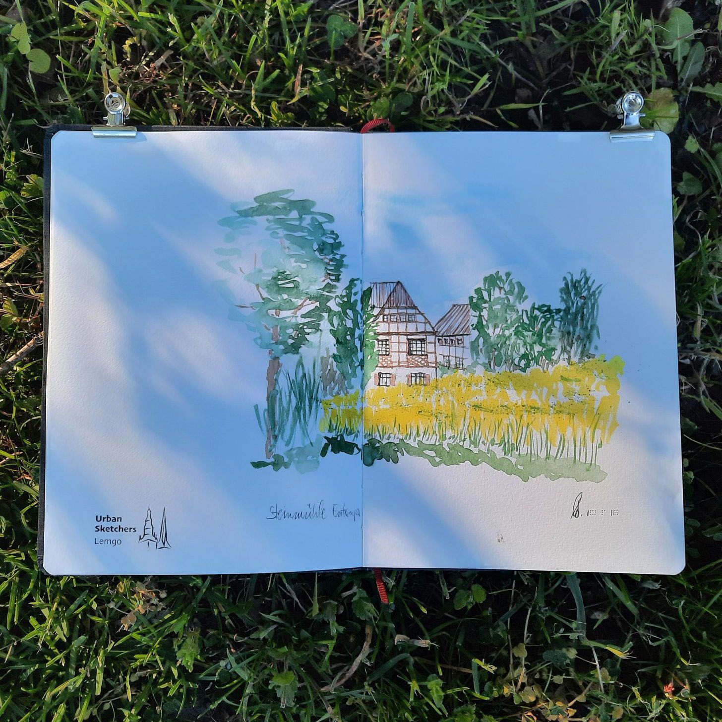 Aufgeschlagenes Skizzenbuch, das auf einer Grasfläche liegt. Aquarelliert ist ein gelbes Rapsfeld, dahinter ein Fachwerkhaus und Bäume.