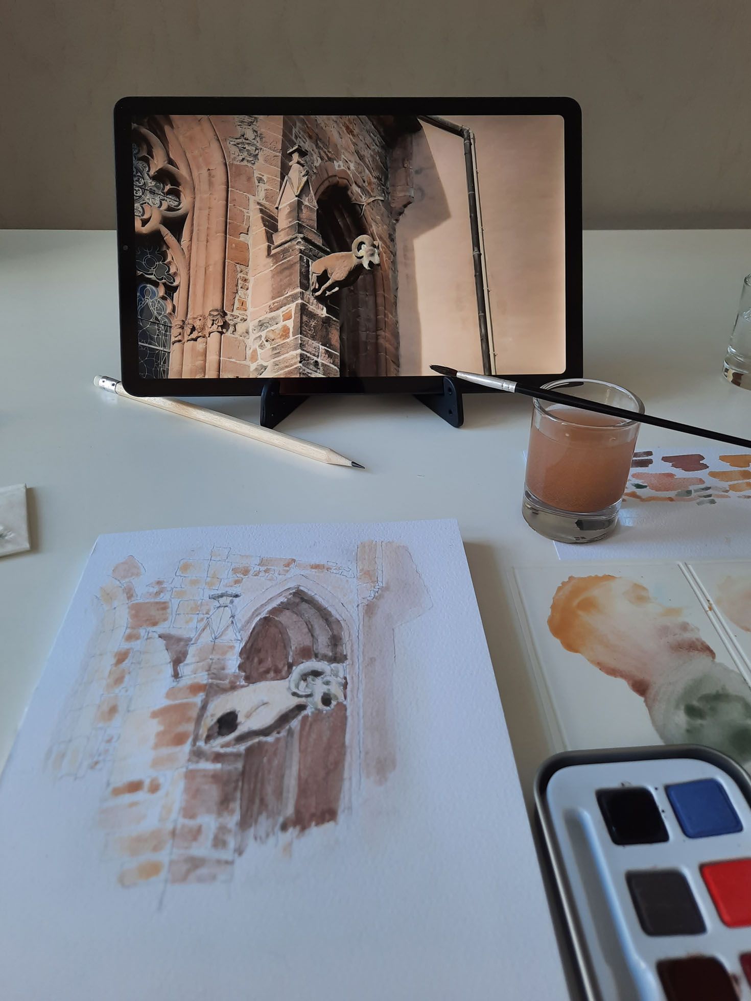 Im Vordergrund ein Skizzenblock mit einer Zeichnung. Dahinter ein senkrecht stehendes Tablet mit einer Fotovorlage.