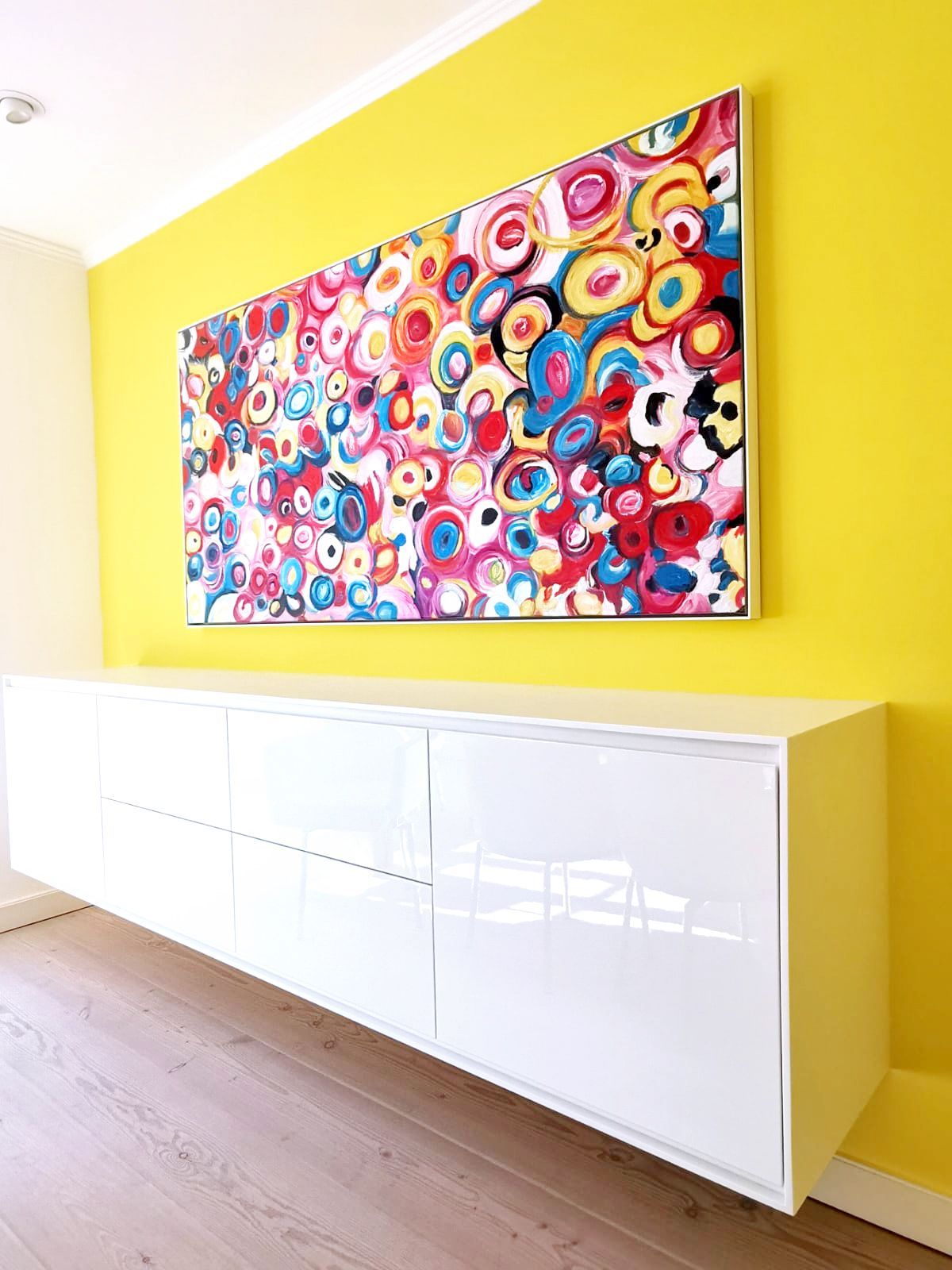 gelbe Akzentwand mit buntem Kunstgemälde über einem weißen hängenden Hochglanz Sideboard