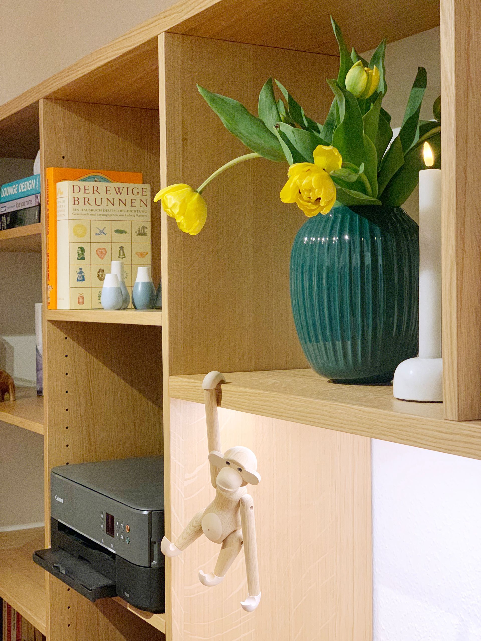 Regalsystem mit Schrankwand, integriertem Schreibtisch, LED-Leiste, Regalfächern, Schubladen und einer Vase mit Tulpen.