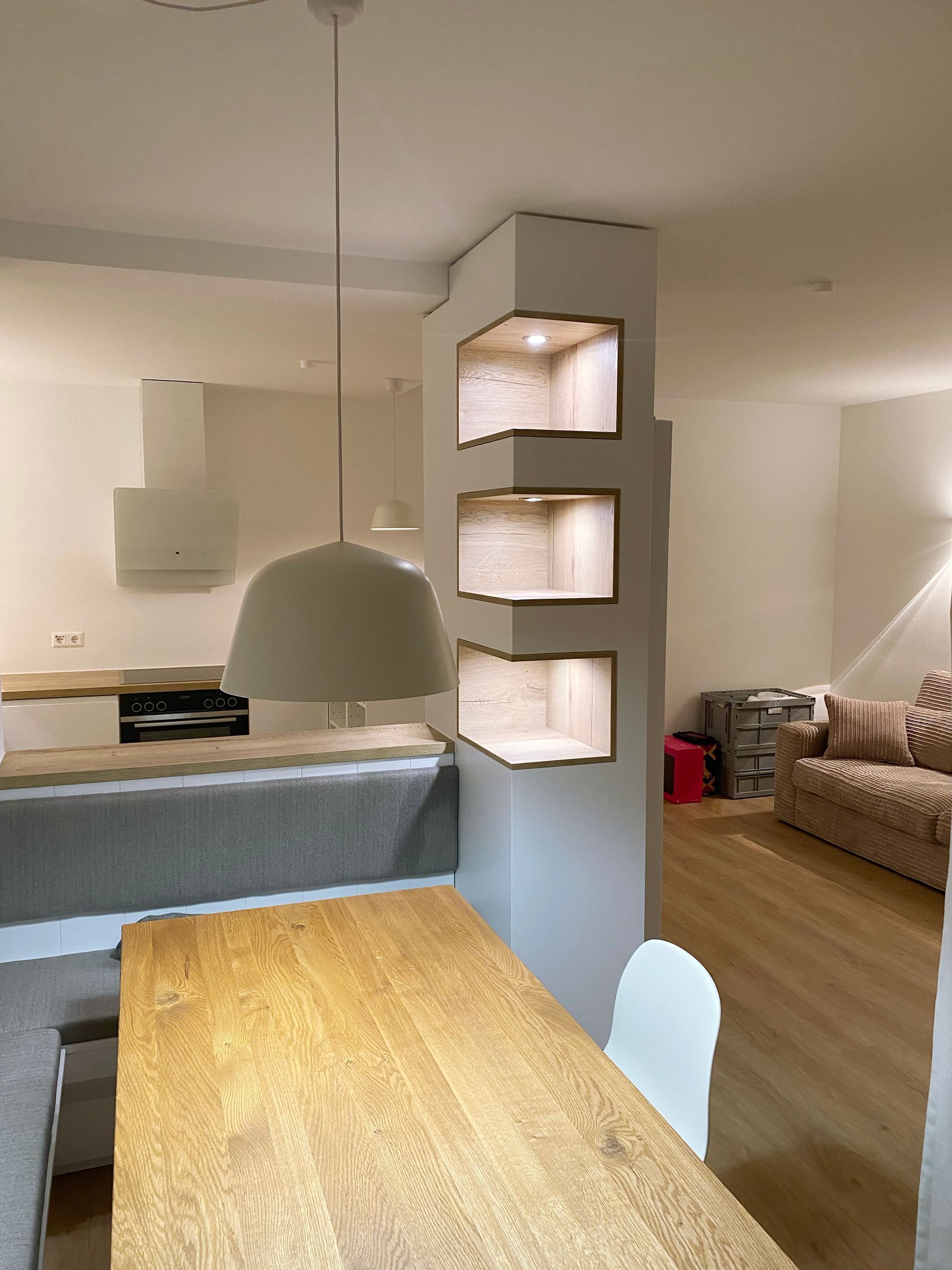 Essecke mit Sitzbank, Esstisch, Regalsäule mit LED-Strahlern und offener Küche in heller Ferienwohnung.