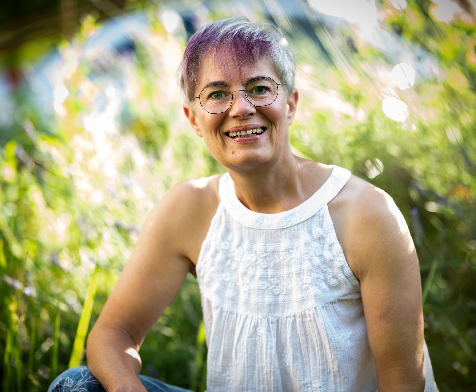 Eine Frau mit kurzem grauem Haar und Brille lächelt im sonnenbeschienenen Laub.