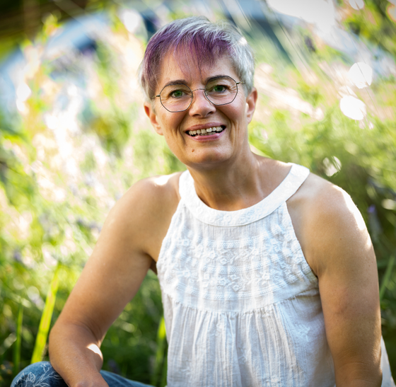 Eine Frau mit kurzem grauem Haar und Brille lächelt im sonnenbeschienenen Laub.