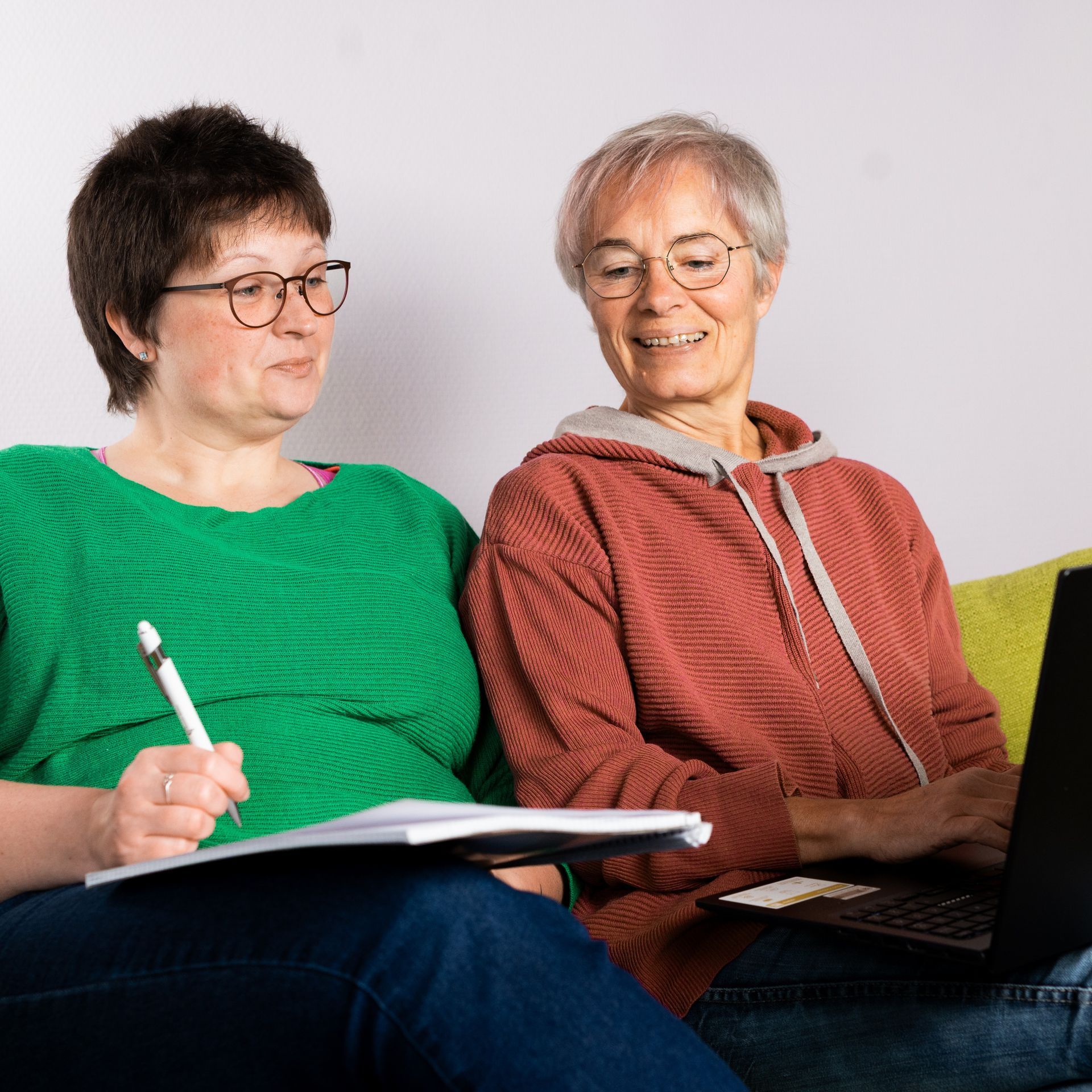 Zwei Frauen sitzen beieinander, die eine benutzt einen Laptop, die andere schreibt in ein Notizbuch, beide tragen Brillen.